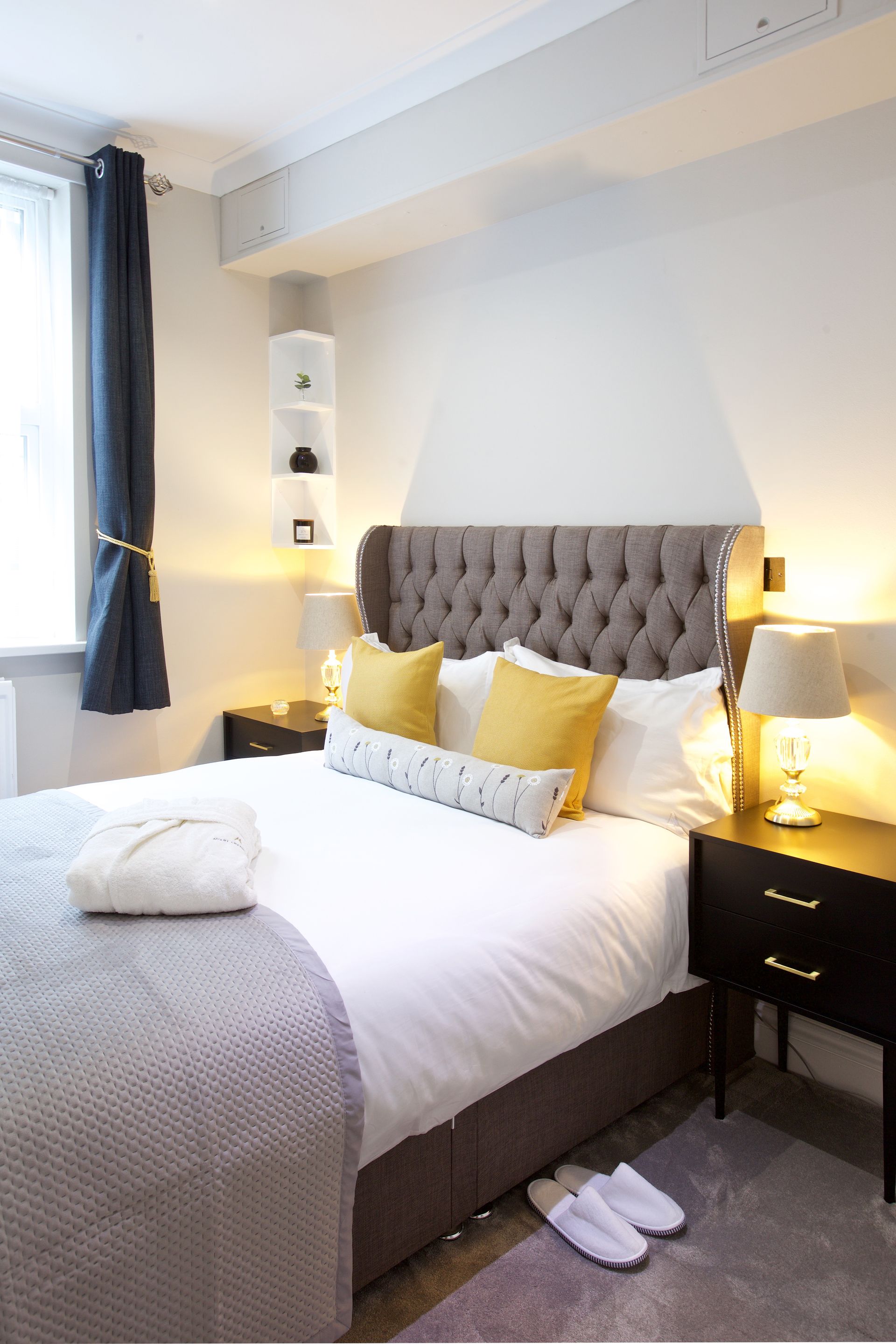 Bright bedroom with tufted gray headboard, white bedding, yellow pillows, and bedside lamps.