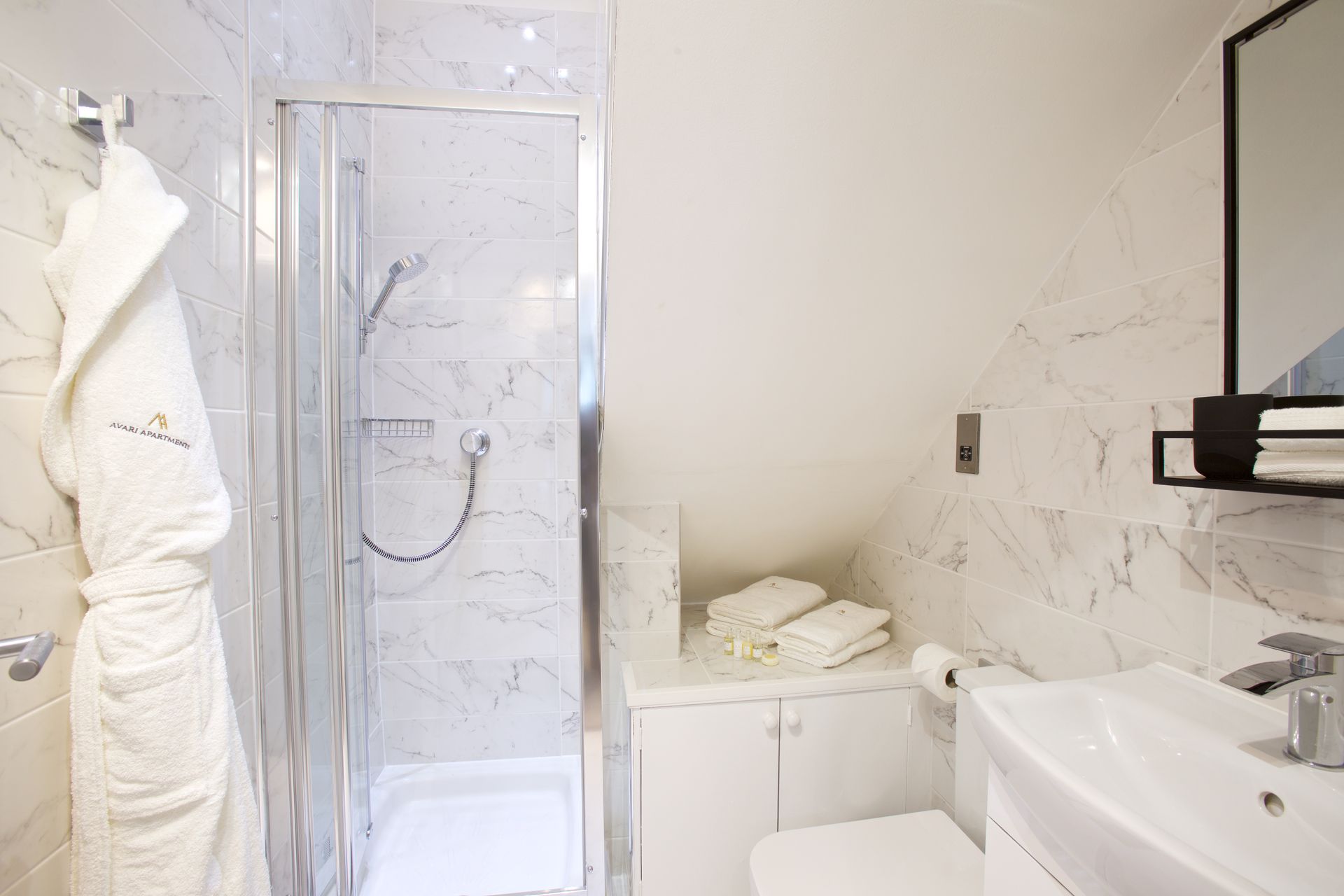 Small white bathroom with shower, sink, mirror, and folded towels on a cabinet.