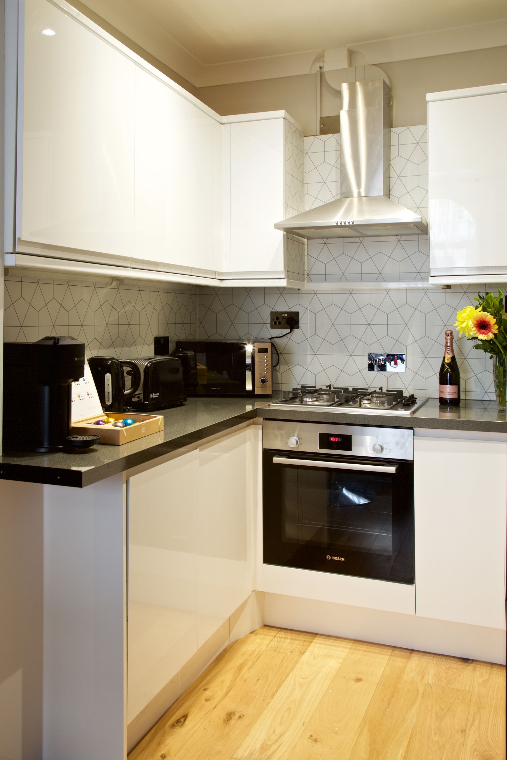 Compact white kitchen with L-shaped counters, oven, range hood, and a vase of sunflowers.
