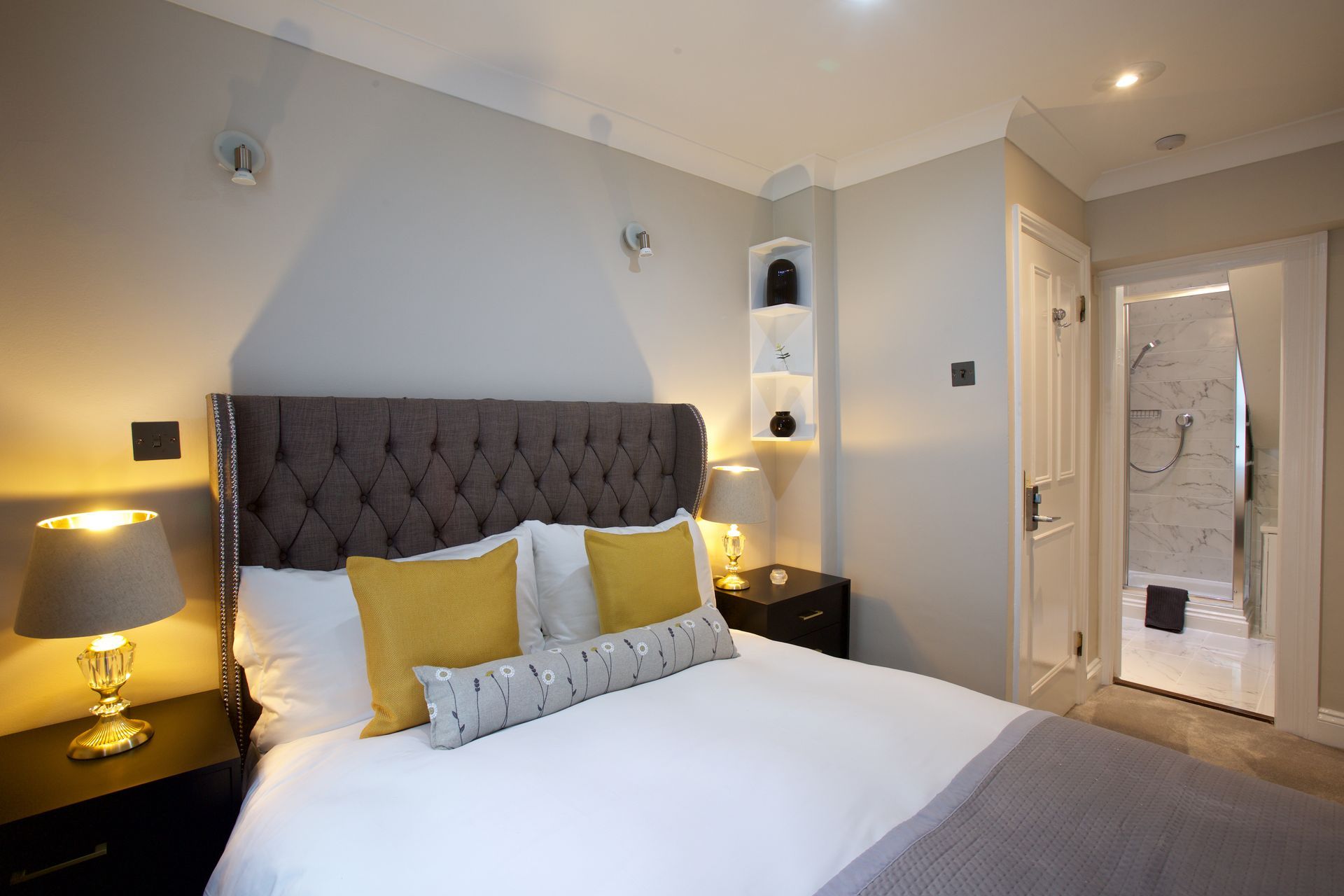 Modern bedroom with tufted gray headboard, white bedding, yellow pillows, and ensuite bathroom door