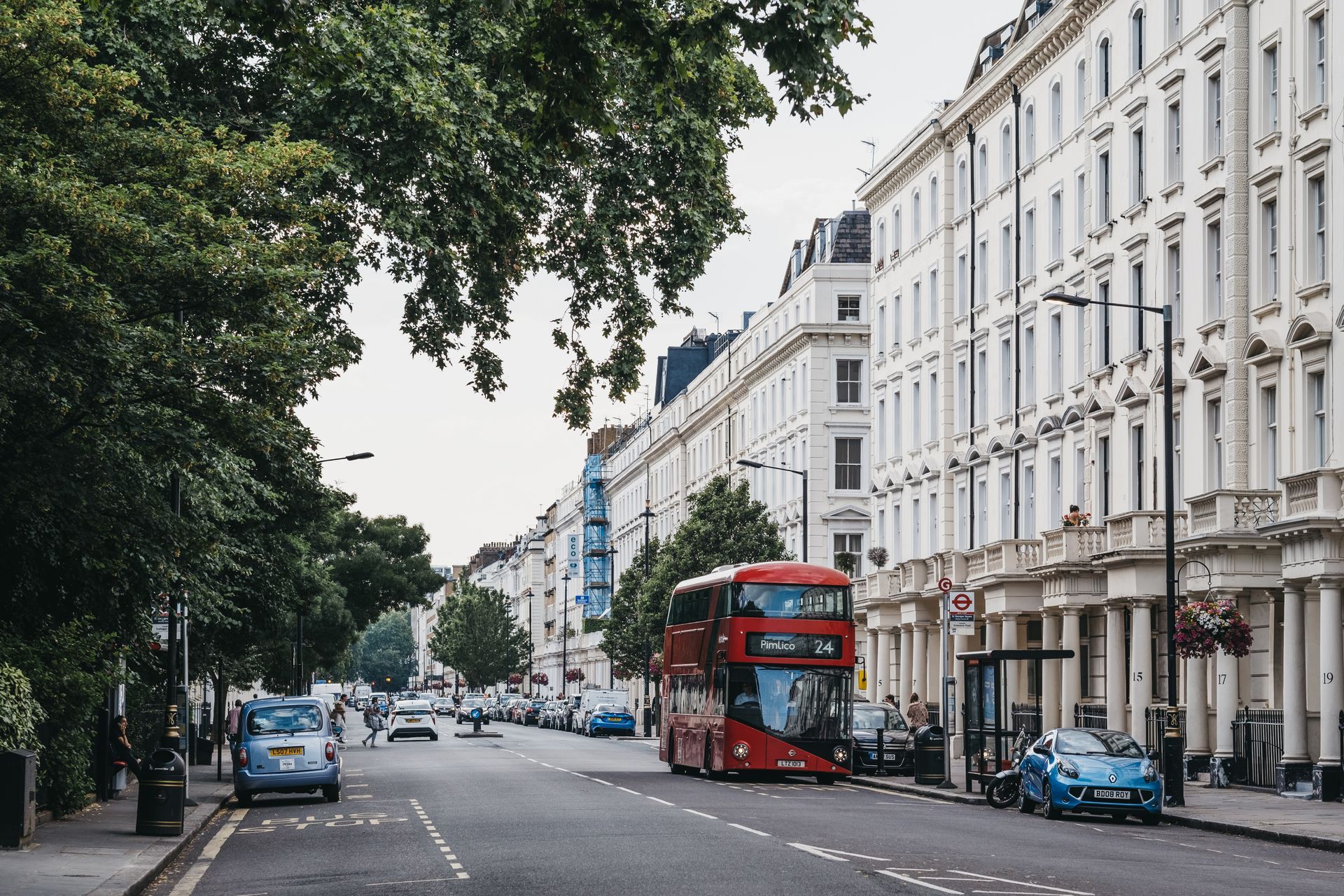 Tree-lined city street with a red double-decker bus and parked cars beside white townhouses