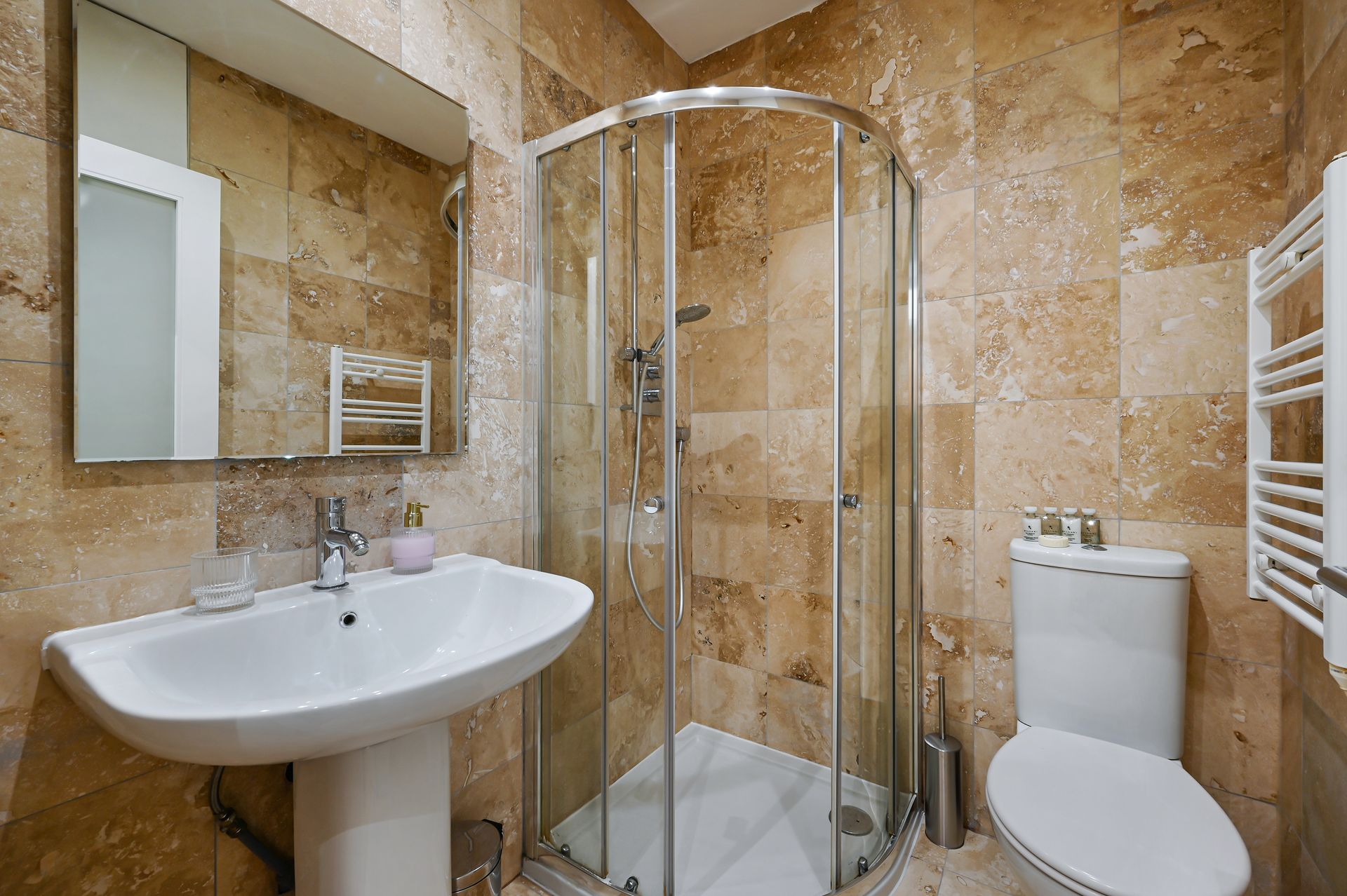 Compact beige-tiled bathroom with sink, shower enclosure, toilet, and towel radiator