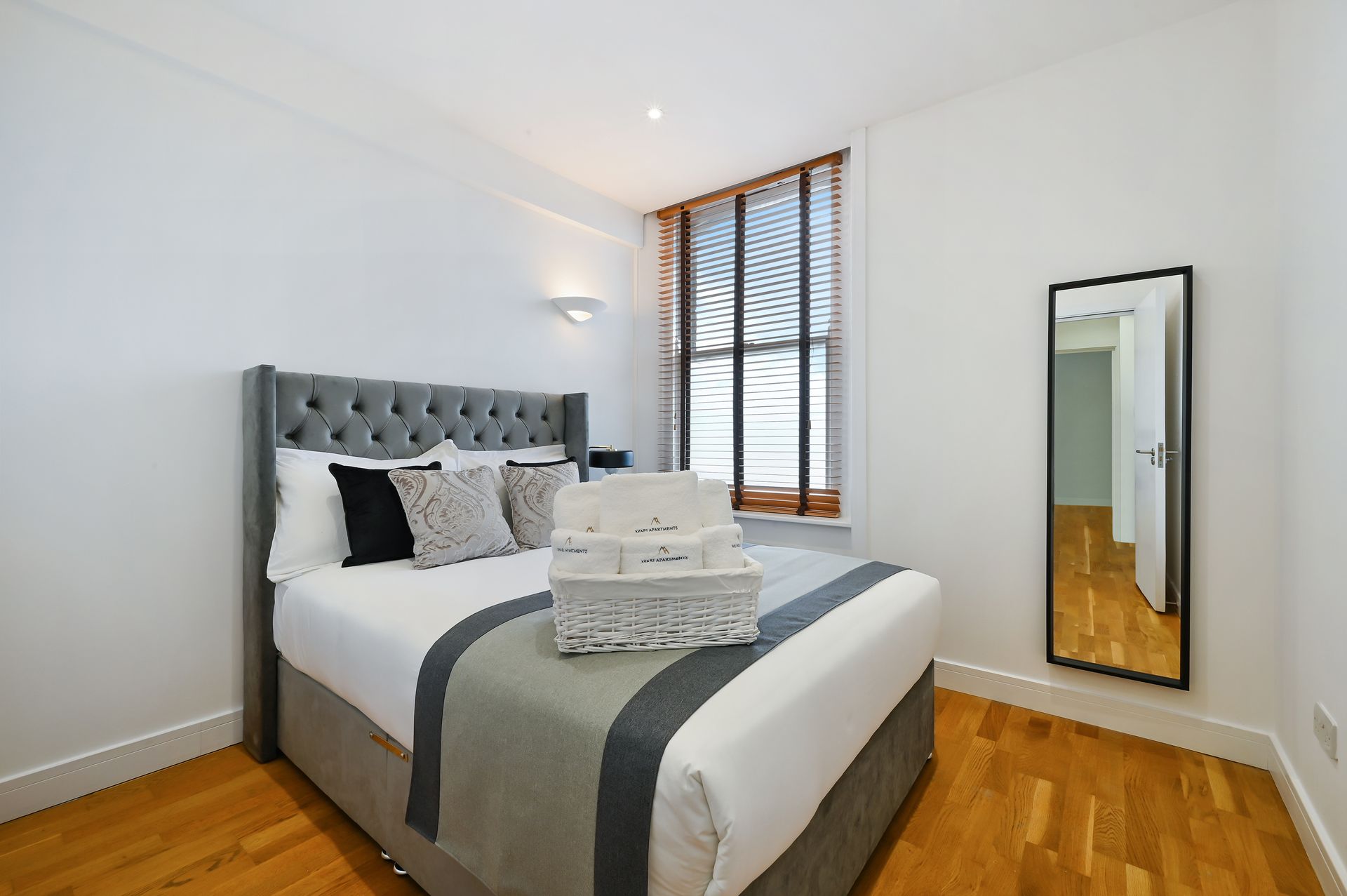 Bright bedroom with gray tufted bed, hardwood floors, striped curtains, and full-length mirror