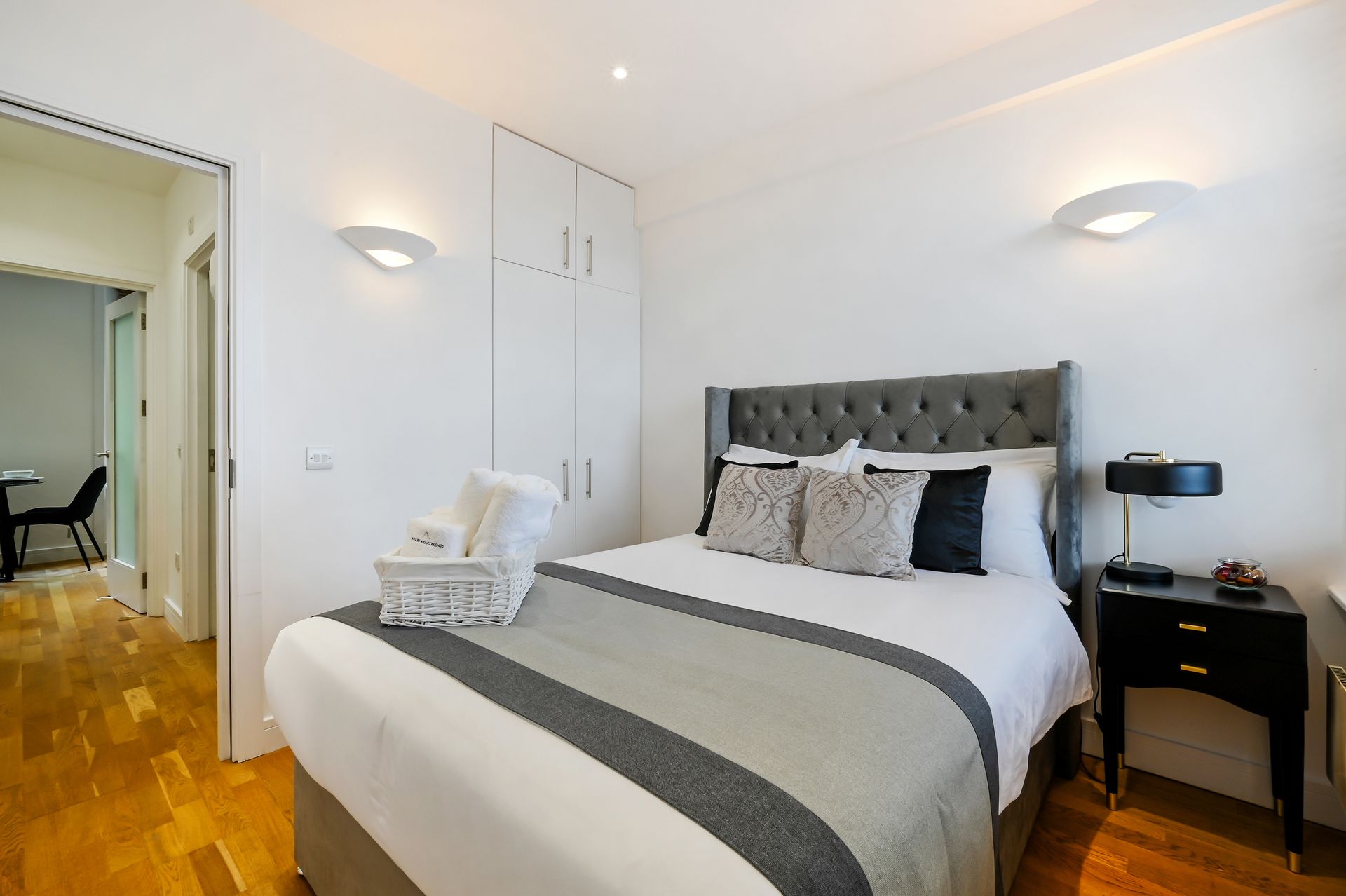 Bright bedroom with a neatly made bed, gray headboard, white walls, and hardwood floor