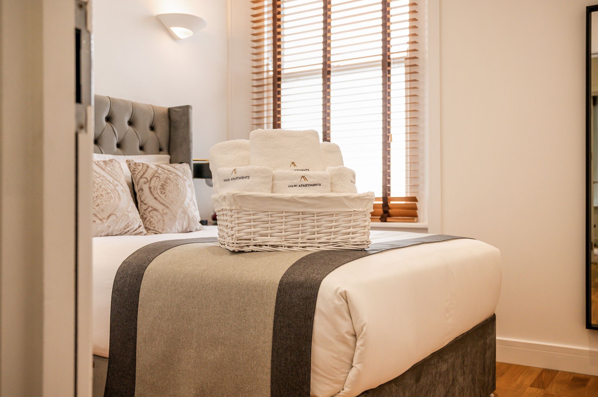 Neatly made bedroom with a gray tufted bed, folded towels, and a window with blinds and wood shutters