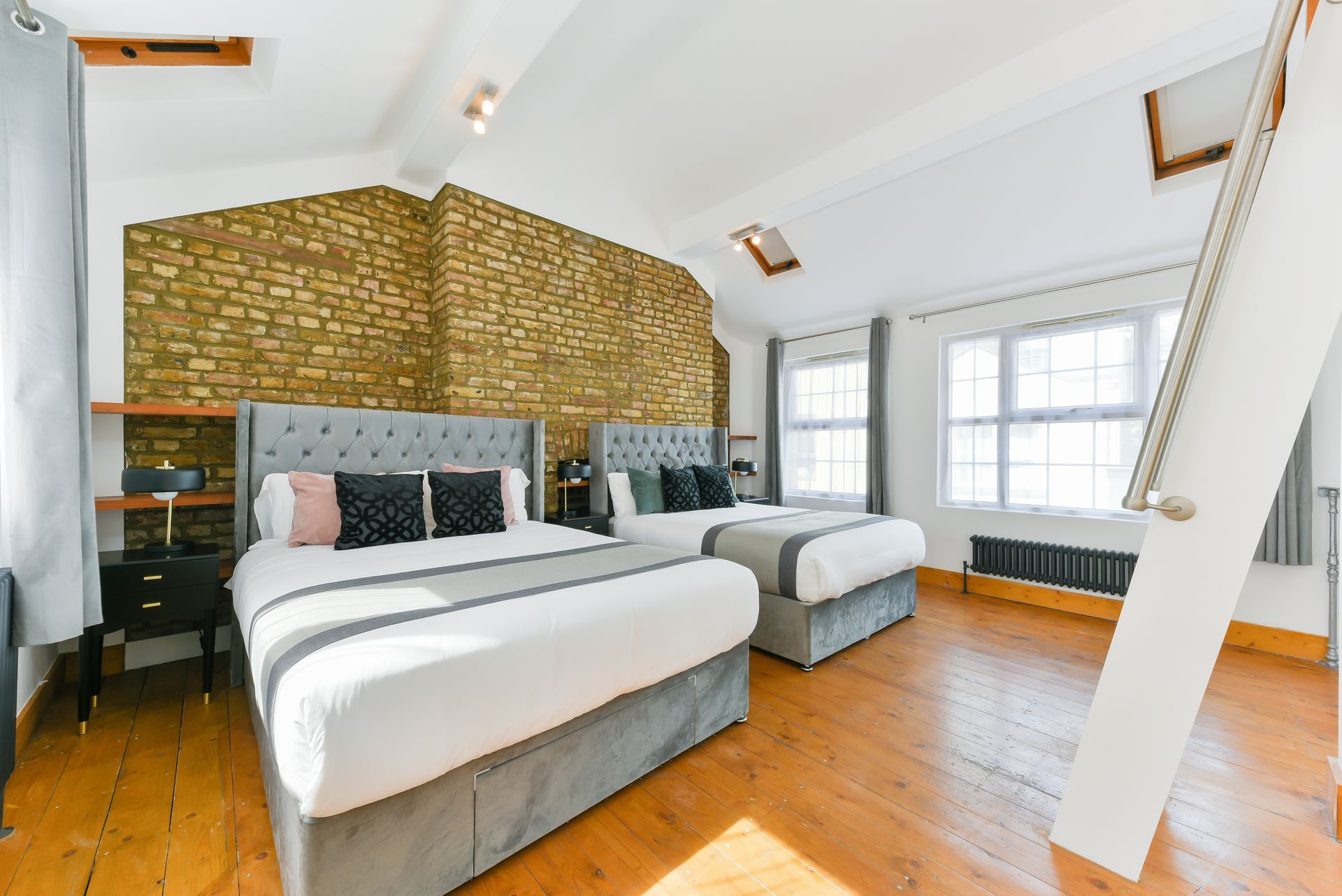 Bright bedroom with two beds, wood floor, patterned brick wall, and large windows with white blinds