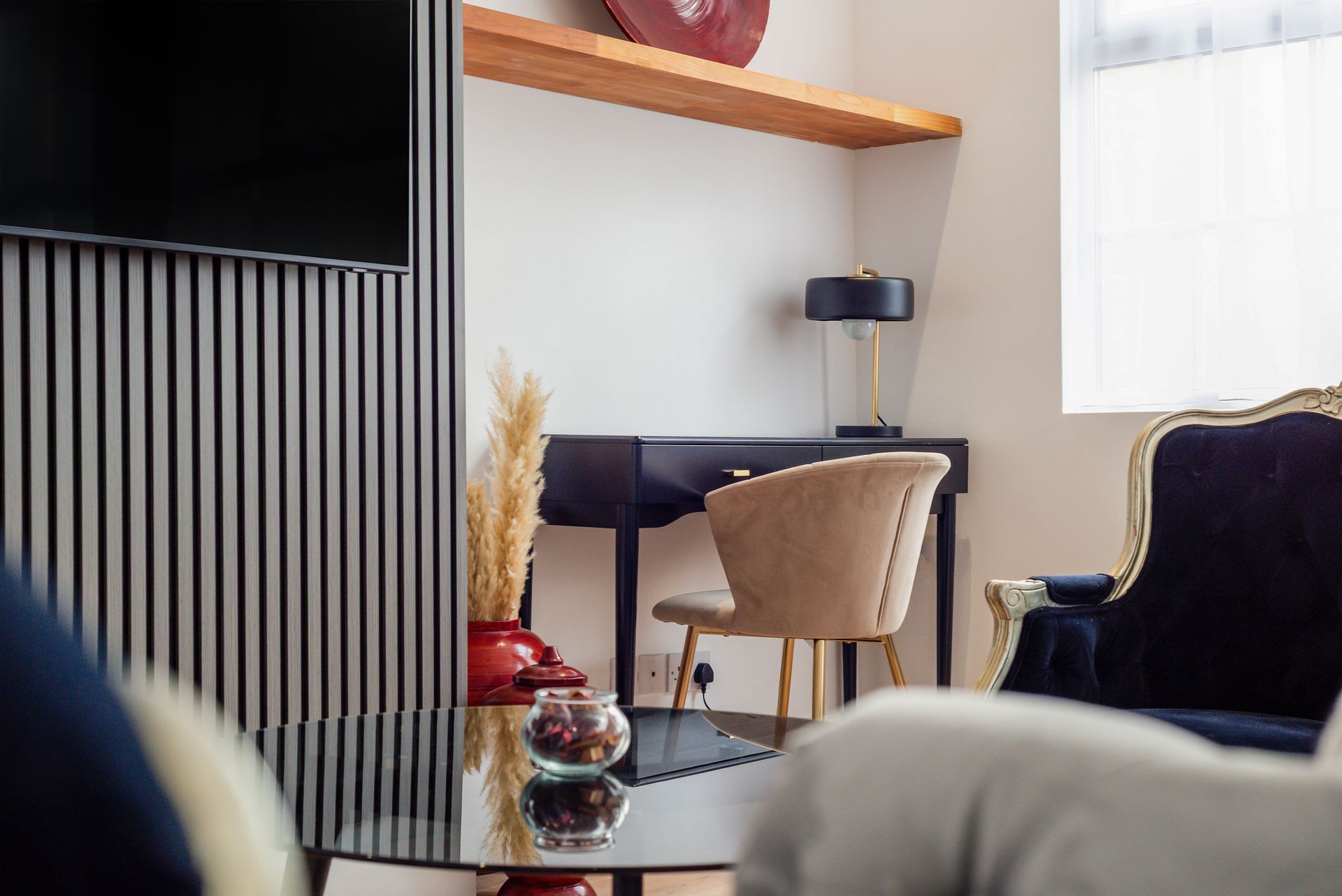 Cozy living room with a desk, beige chair, dark armchair, and glass coffee table near a window.