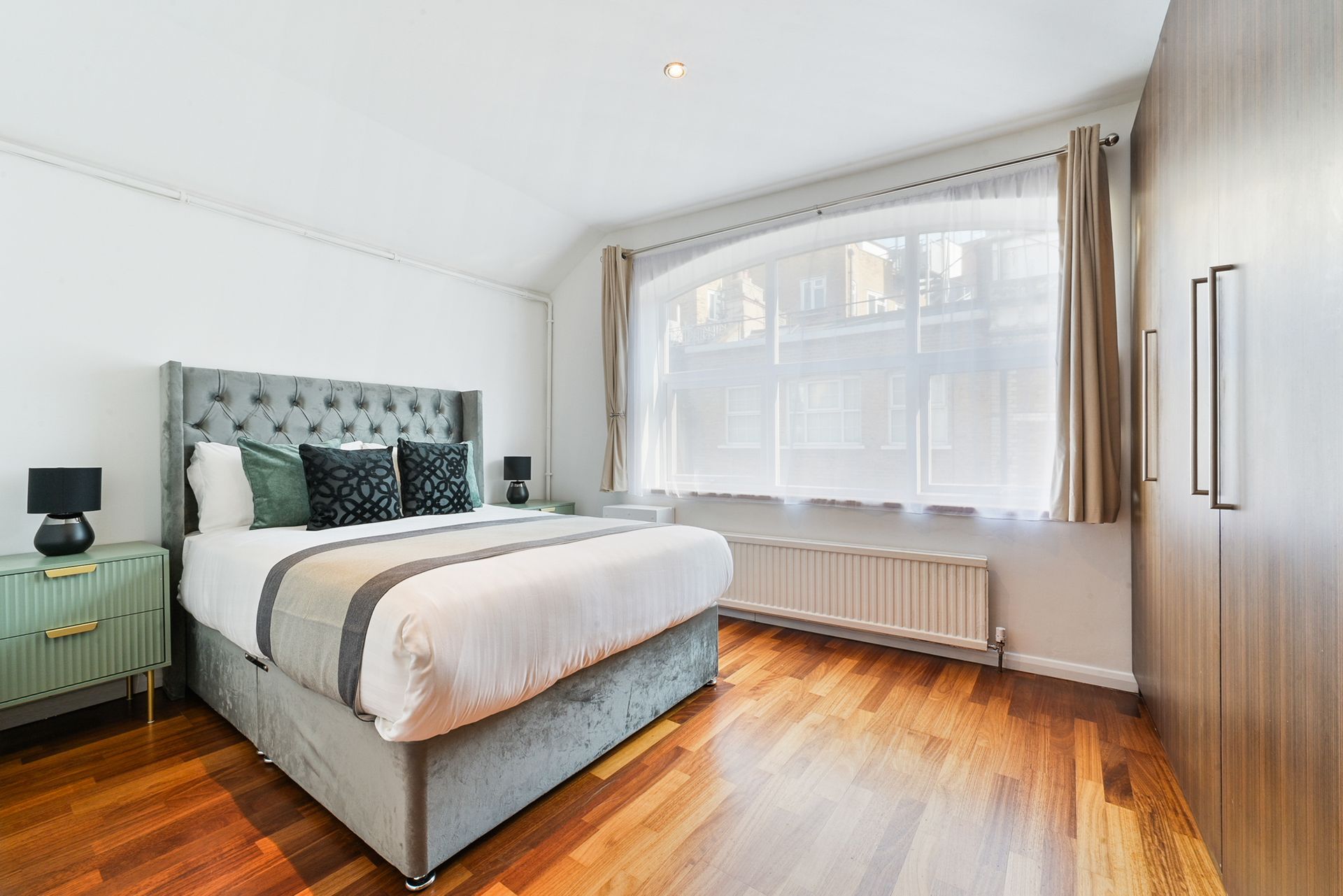 Bright bedroom with teal upholstered bed, white bedding, wooden floor, and a large window with curtains