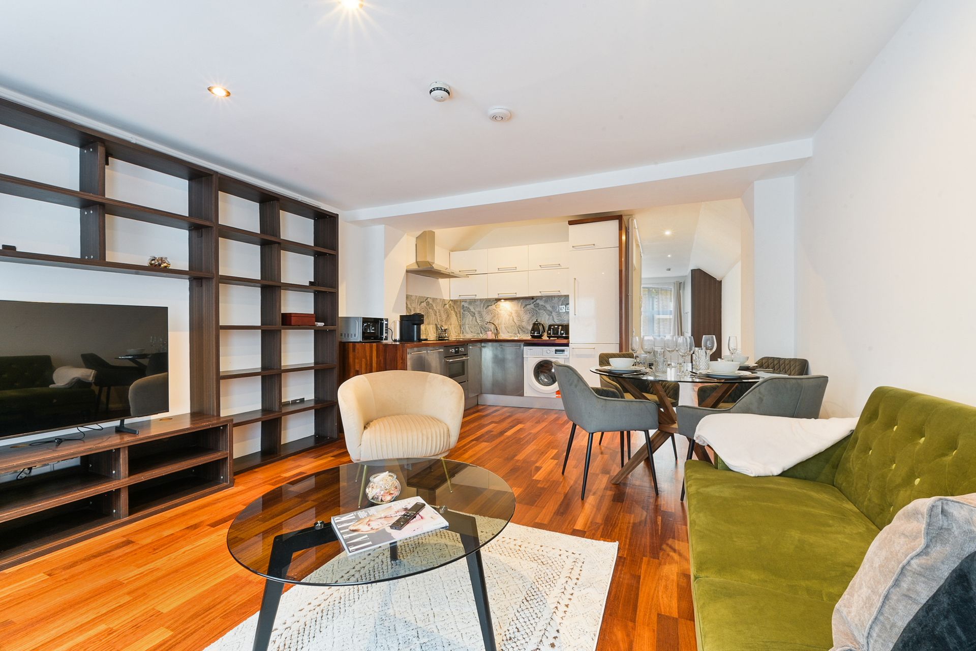 Modern living room with green sofa, black TV stand, dining table, and open kitchen with wood floors.