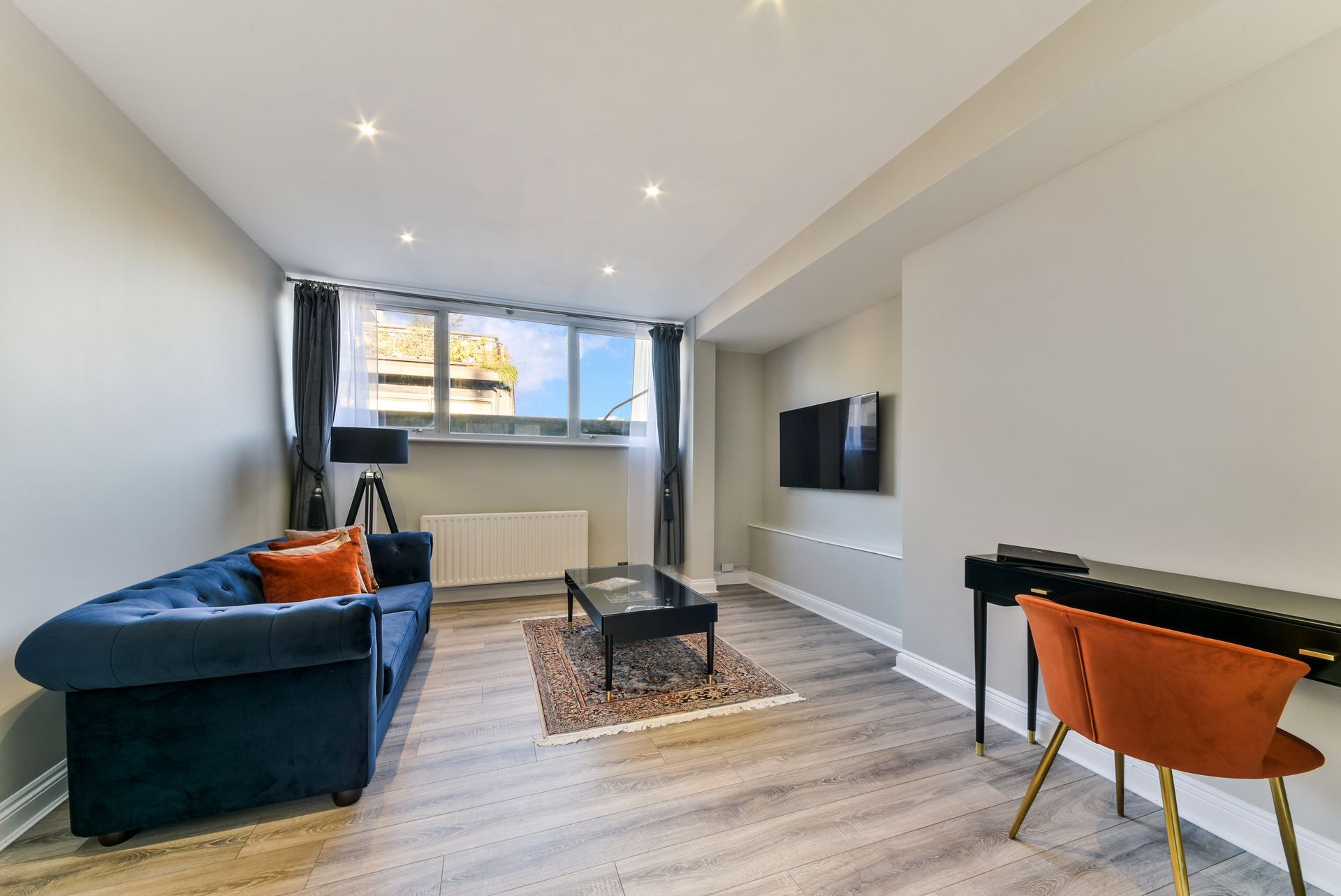 Modern living room with blue sofa, orange chair, wall-mounted TV, coffee table, and large window