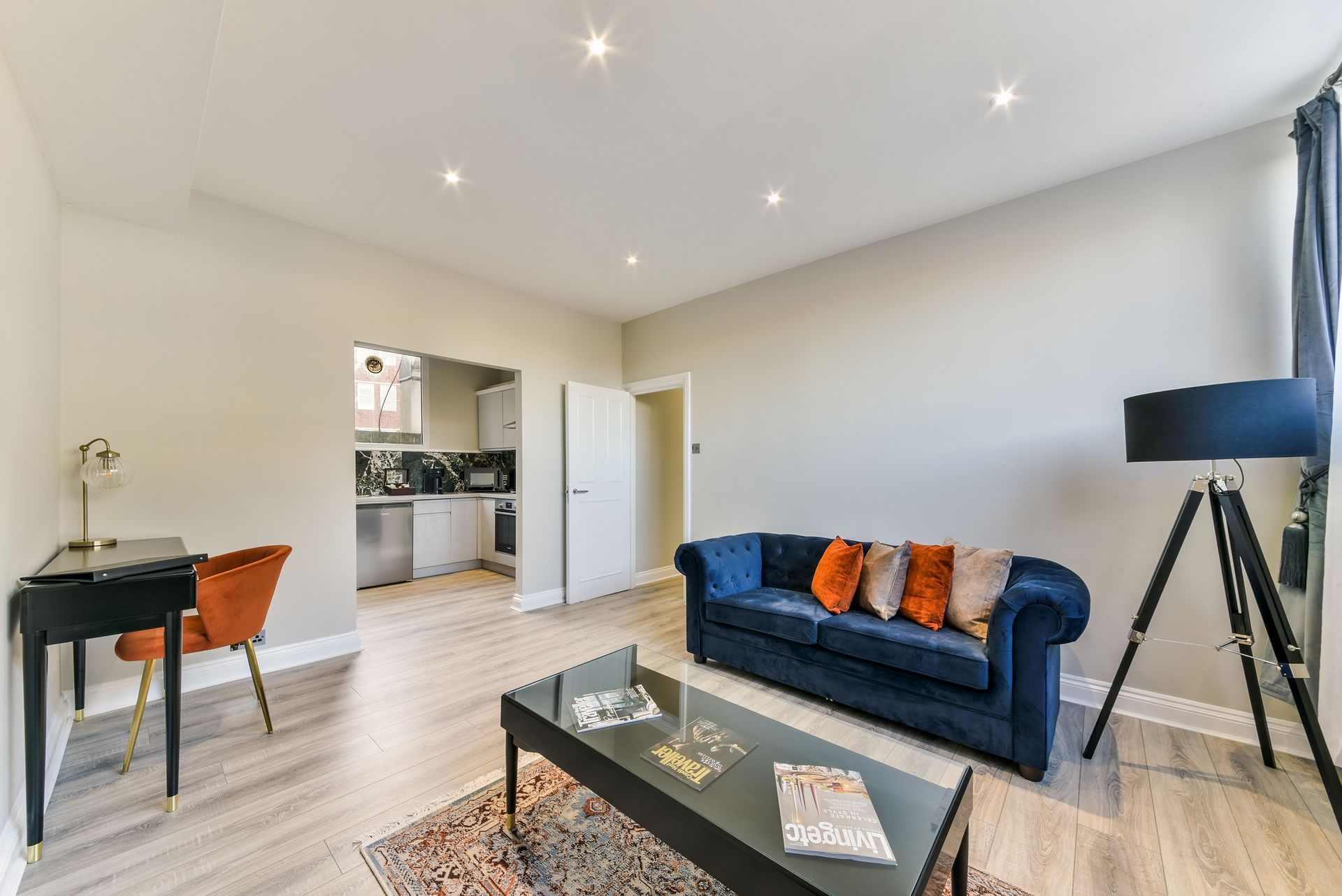 Bright modern living room with blue sofa, glass coffee table, tripod lamp, and open kitchen view