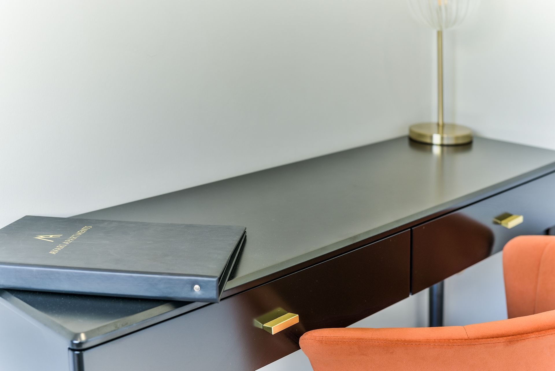 Minimal office desk with gray laptop, brass lamp, and orange chair against a white wall