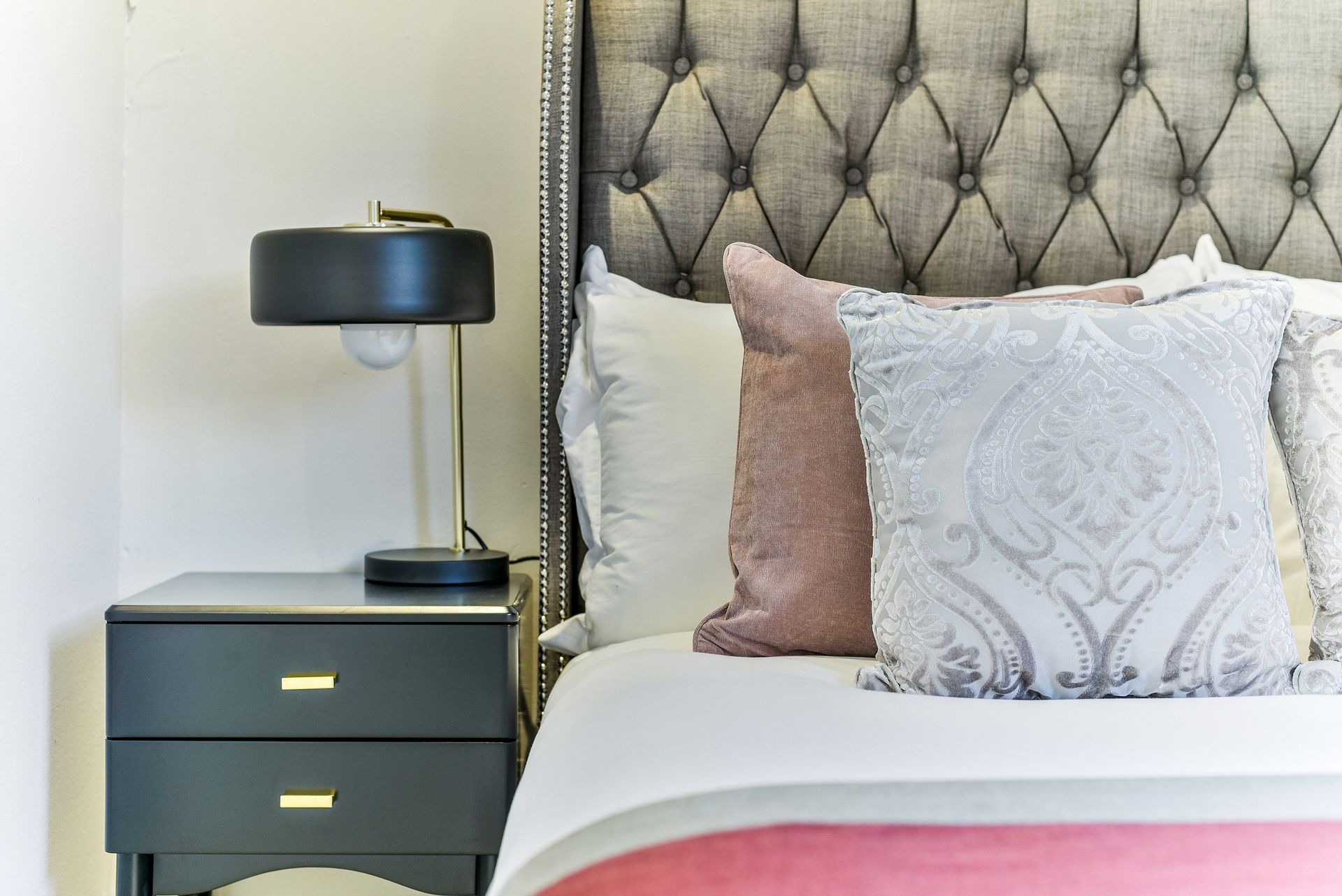 A stylish bedroom with a tufted headboard, gray nightstand, black lamp, and white and pink pillows.
