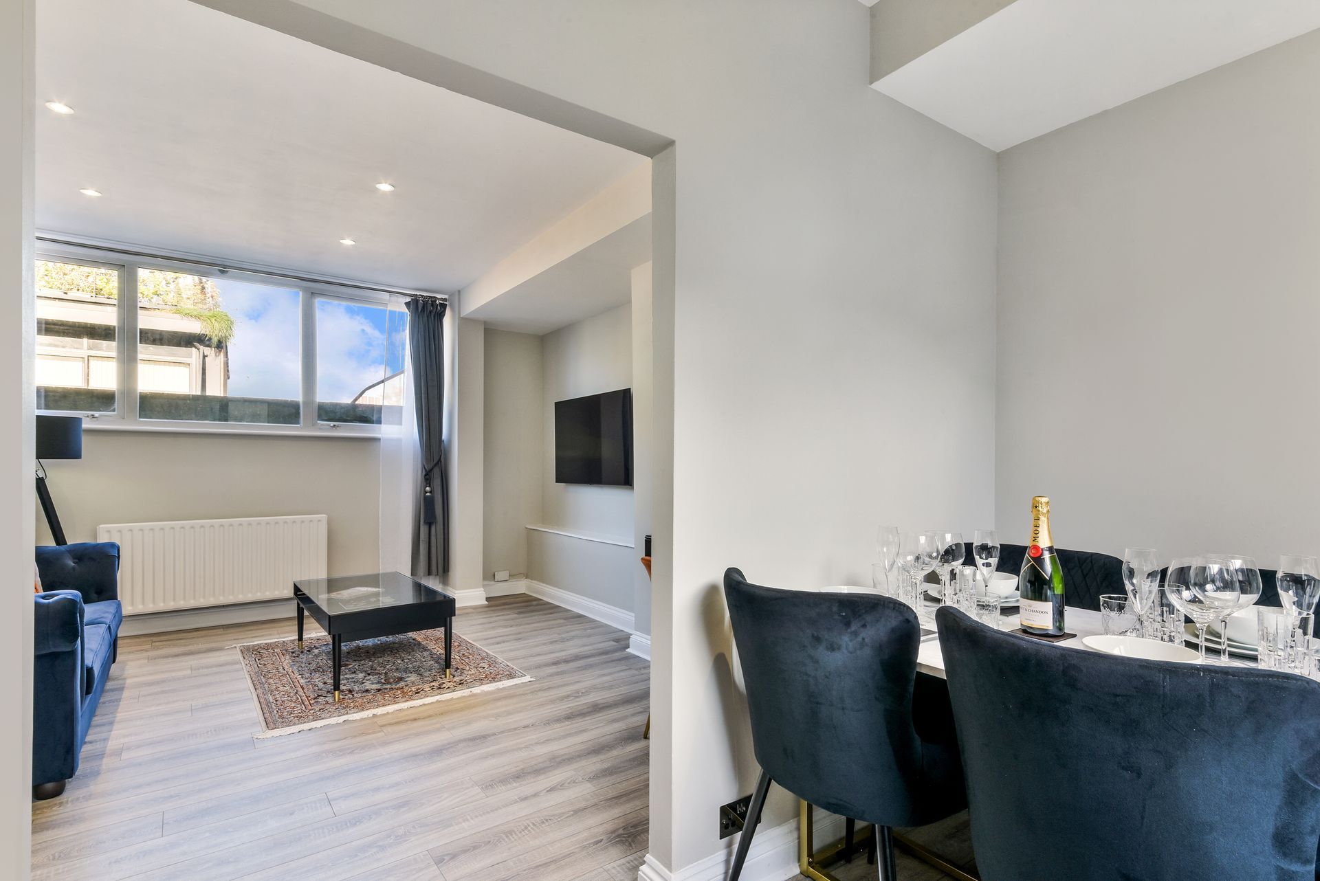 Modern open-plan living and dining room with blue sofa, coffee table, wall TV, and dining table by large windows
