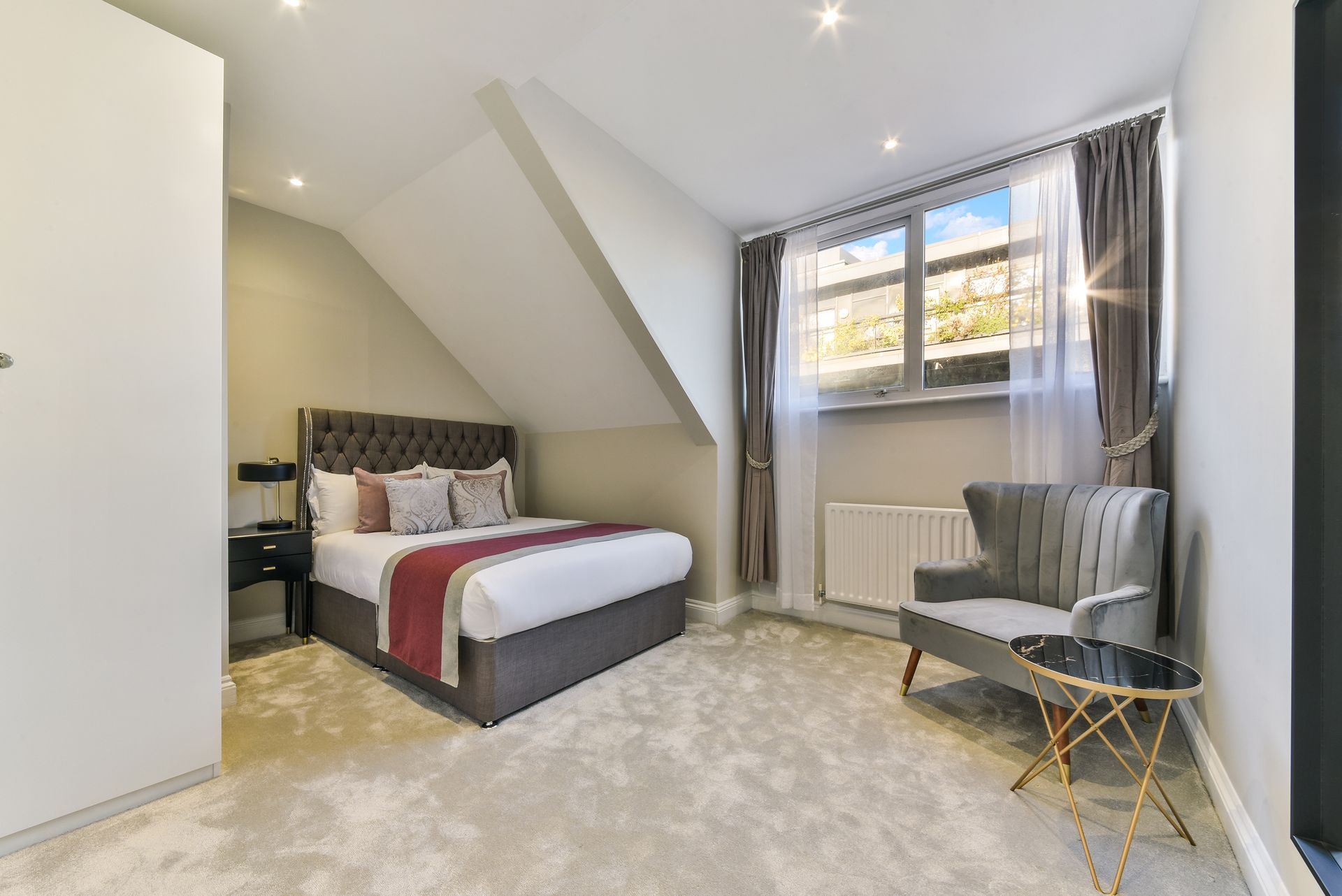 Bright attic bedroom with a bed, armchair, small table, window, and gray carpet.