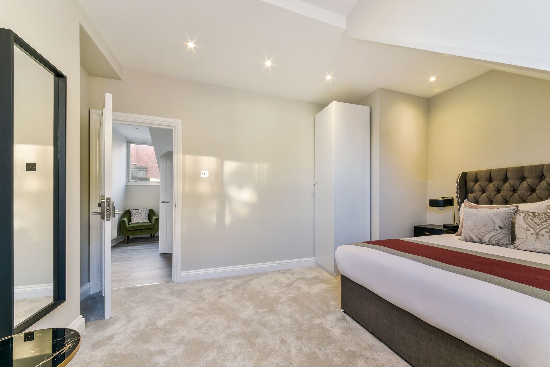Bright modern bedroom with a tufted bed, white wardrobe, full-length mirror, and doorway to a bathroom