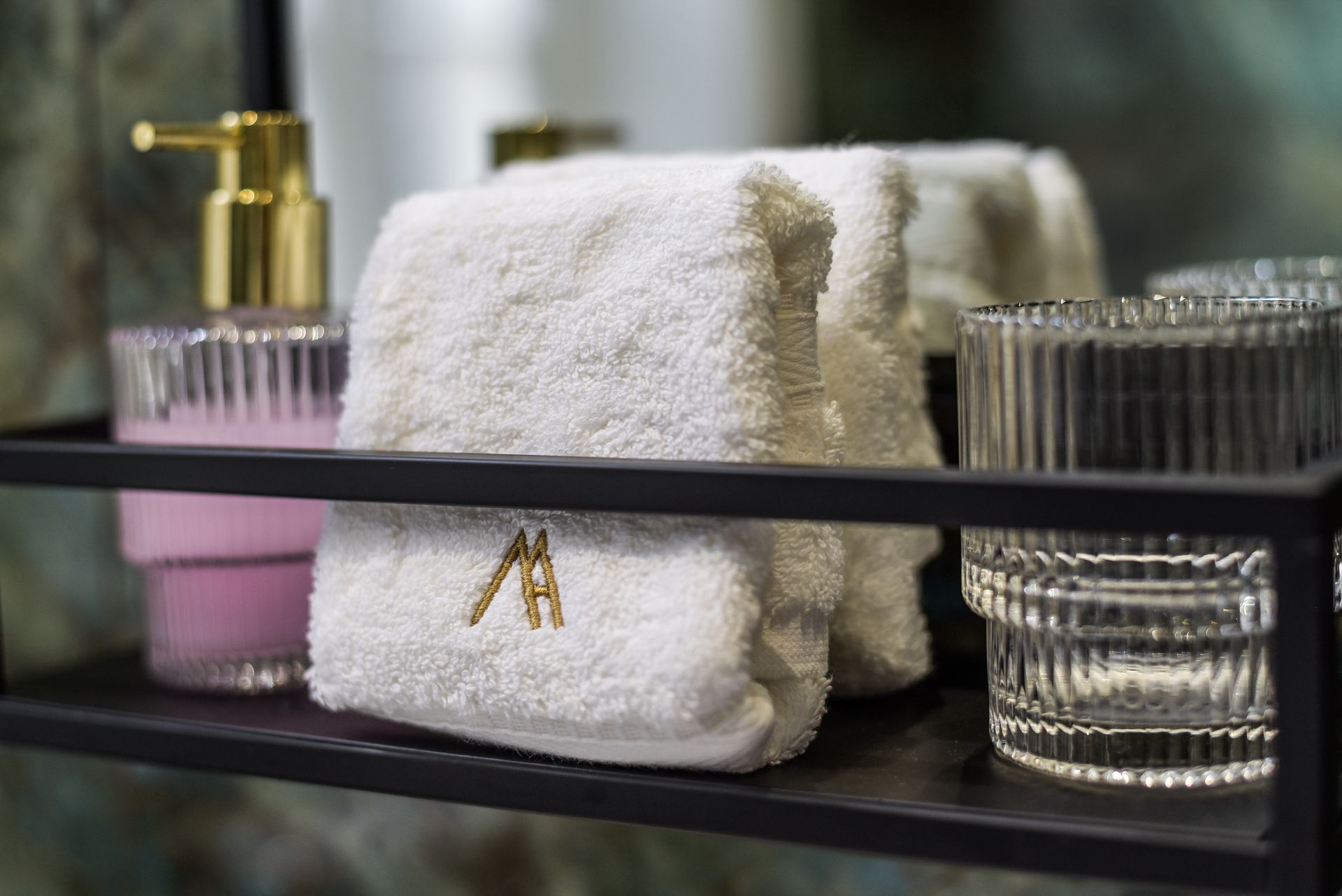 White towels with gold monogram on a bathroom shelf beside glass tumblers and a gold soap dispenser