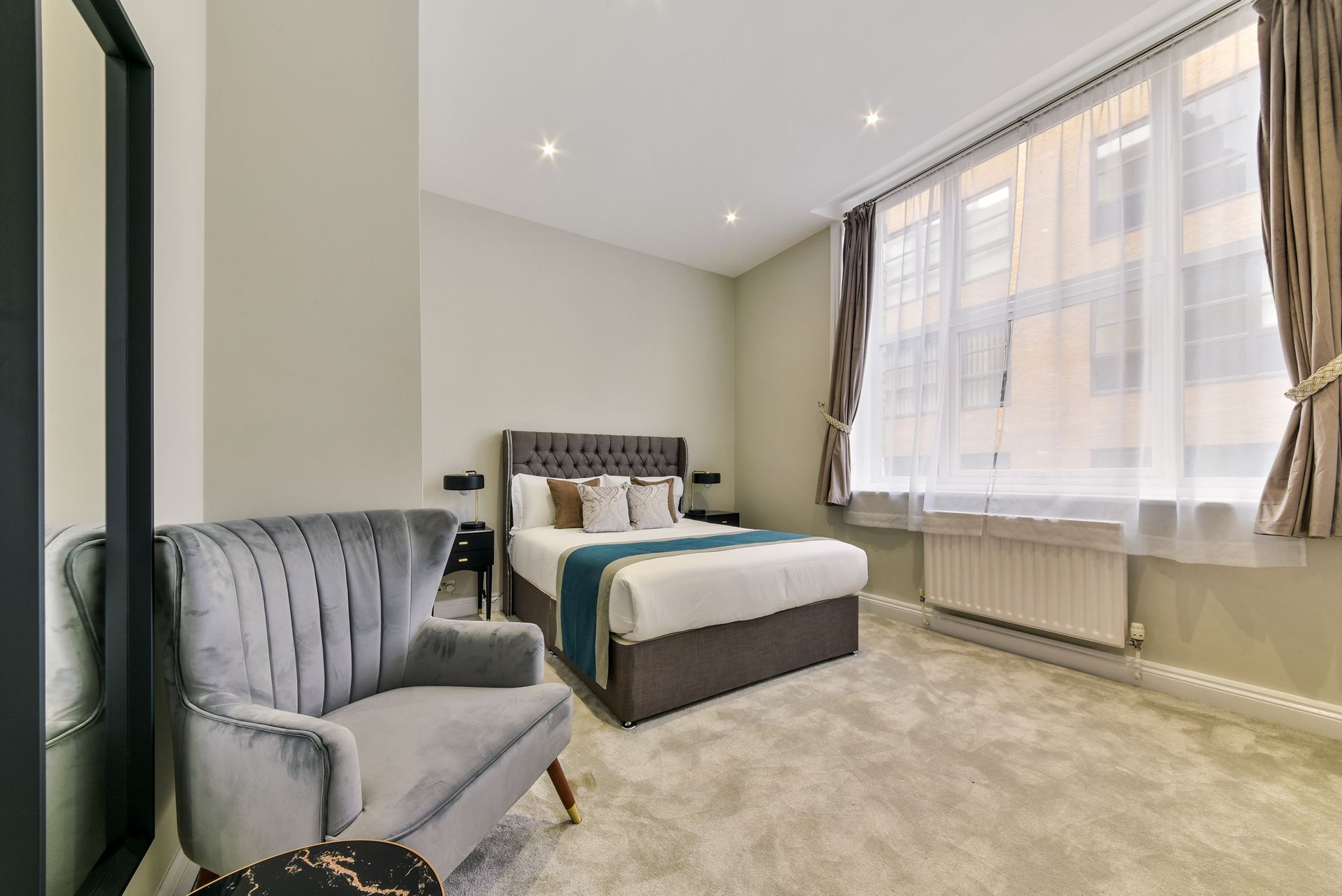 Bright bedroom with bed, gray armchair, carpet, and large window with curtains
