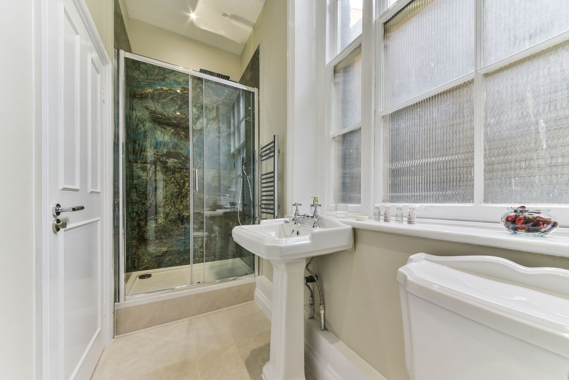 Small white bathroom with glass shower, pedestal sink, toilet, and frosted window
