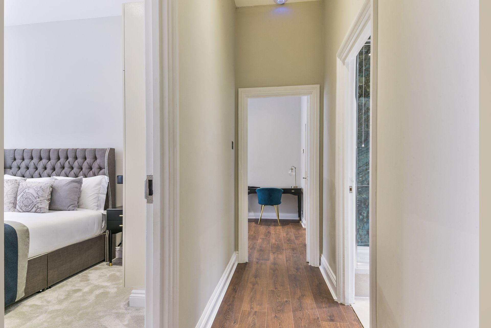 Bright hallway with wood floors, leading to a bedroom with a gray tufted bed and blue chair.