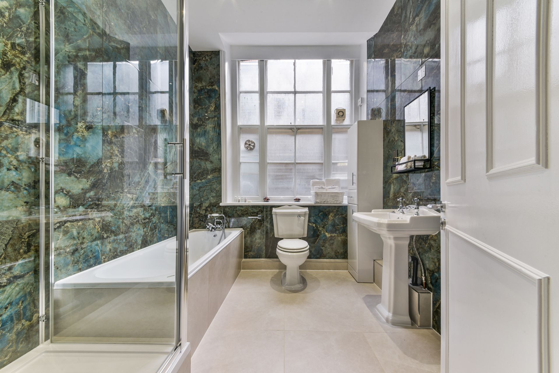 Bright bathroom with a glass shower, bathtub, toilet, pedestal sink, and floral tile walls.