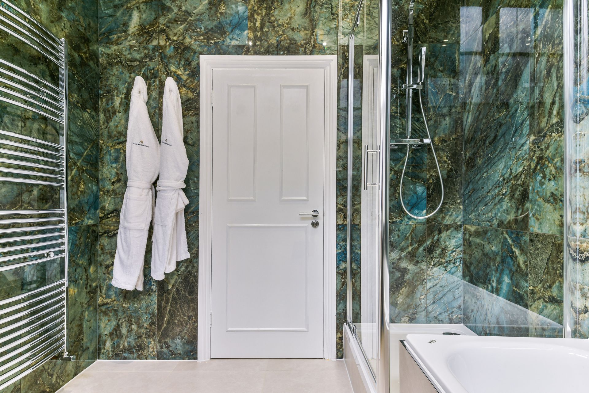 Modern bathroom with white door, marble green walls, hanging robes, and glass shower beside a tub