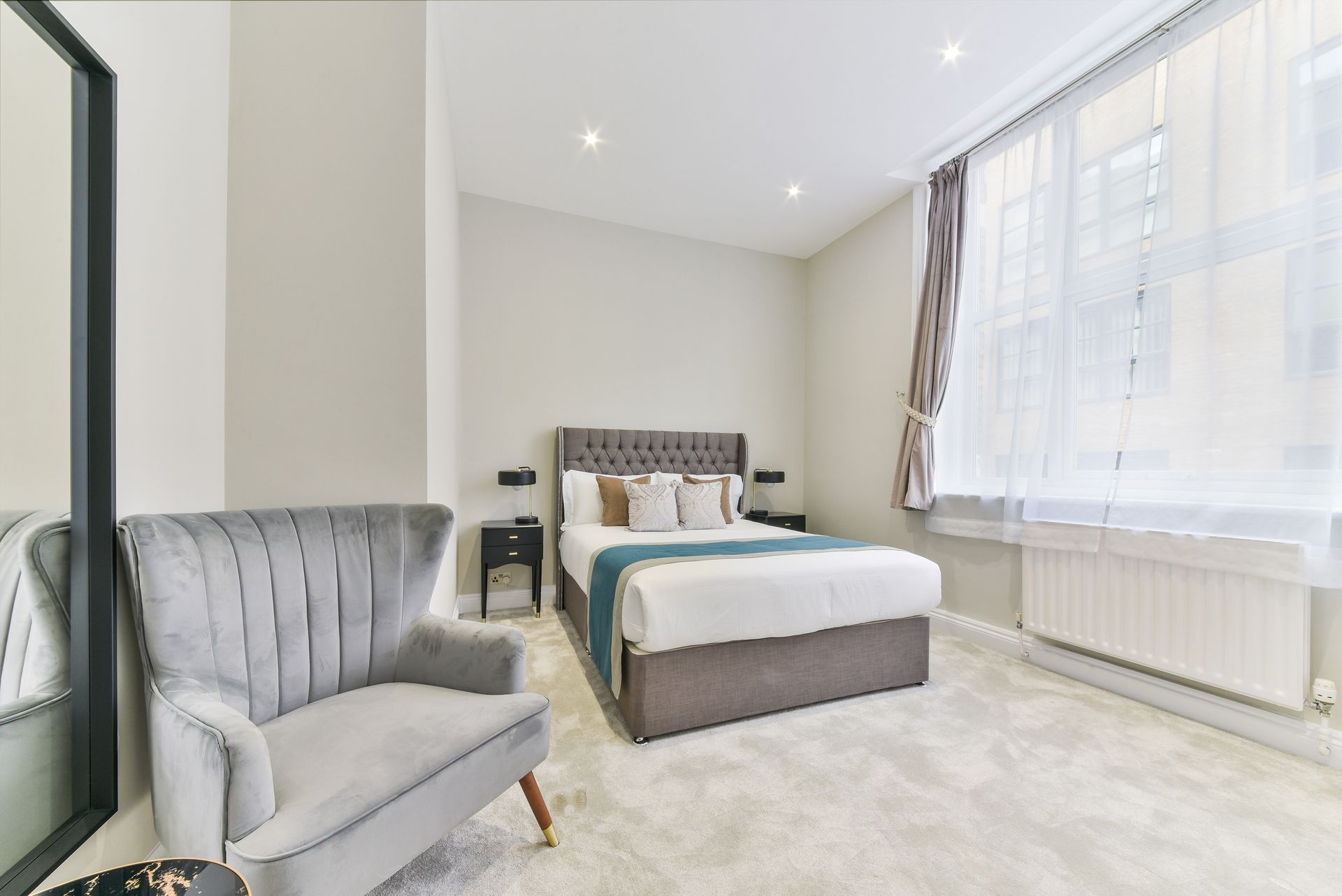 Bright bedroom with a gray upholstered bed, matching chair, and large window with sheer curtains