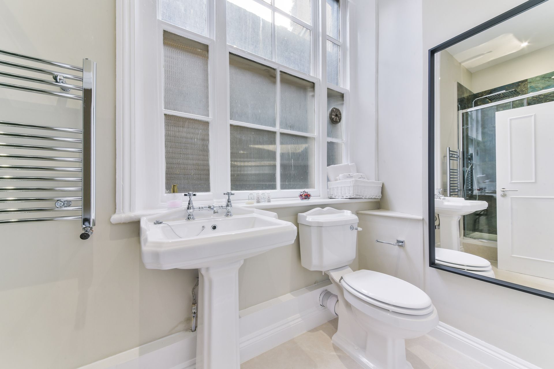 Bright white bathroom with sink, toilet, towel rack, and large mirror beside a window.