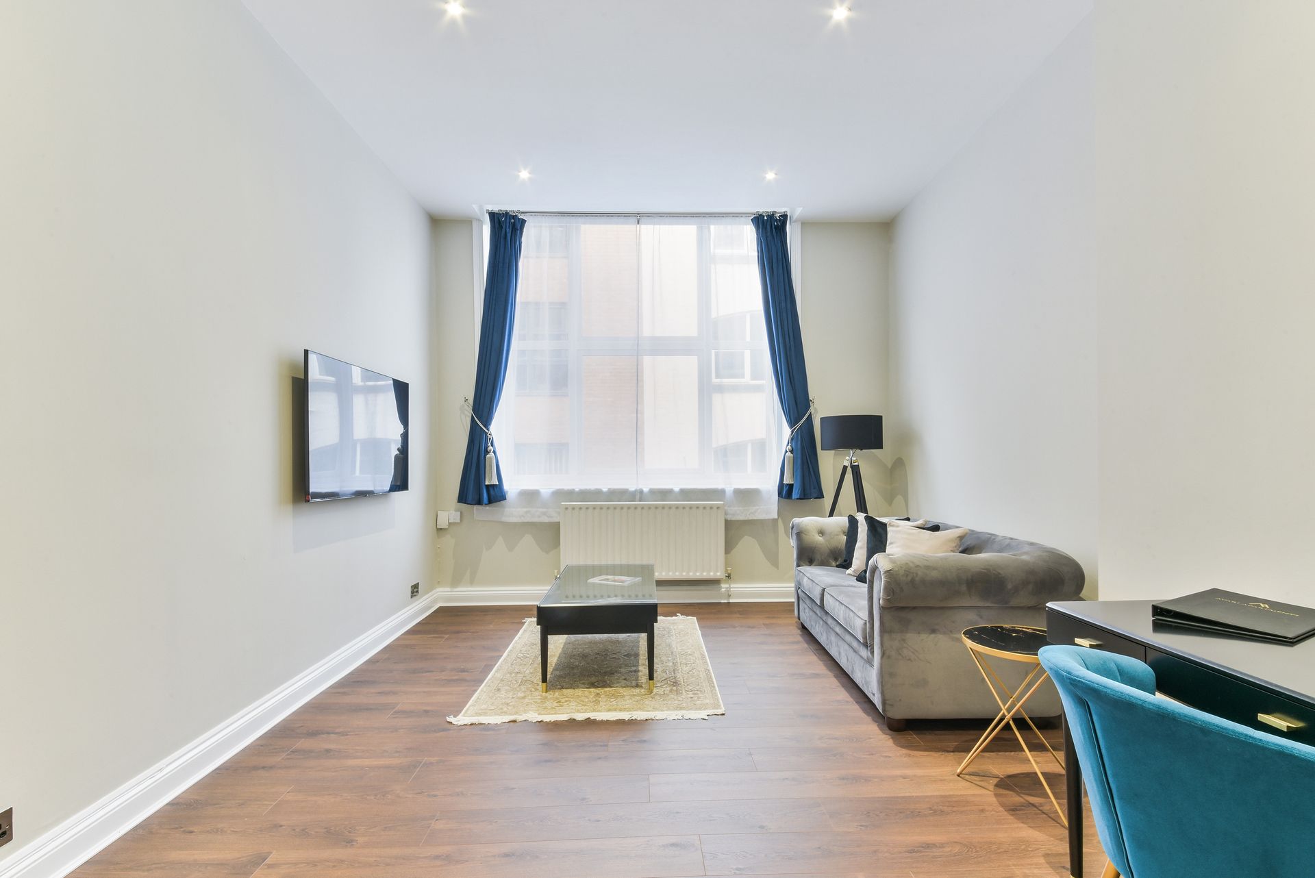 Bright living room with wood floors, blue curtains, gray sofa, TV, and coffee table near a window.