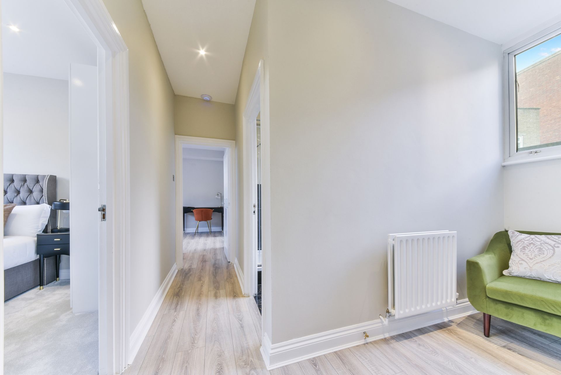 Bright hallway with white walls, wood floor, radiator, and a green chair by a window.