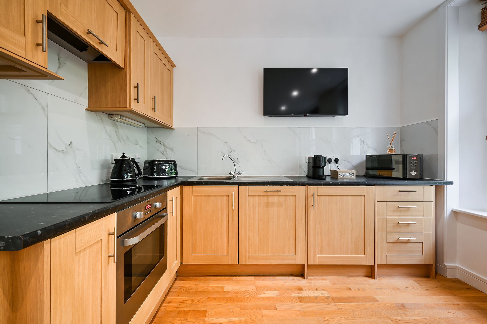 Modern kitchen with light wood cabinets, black countertops, stainless oven, and wall-mounted TV