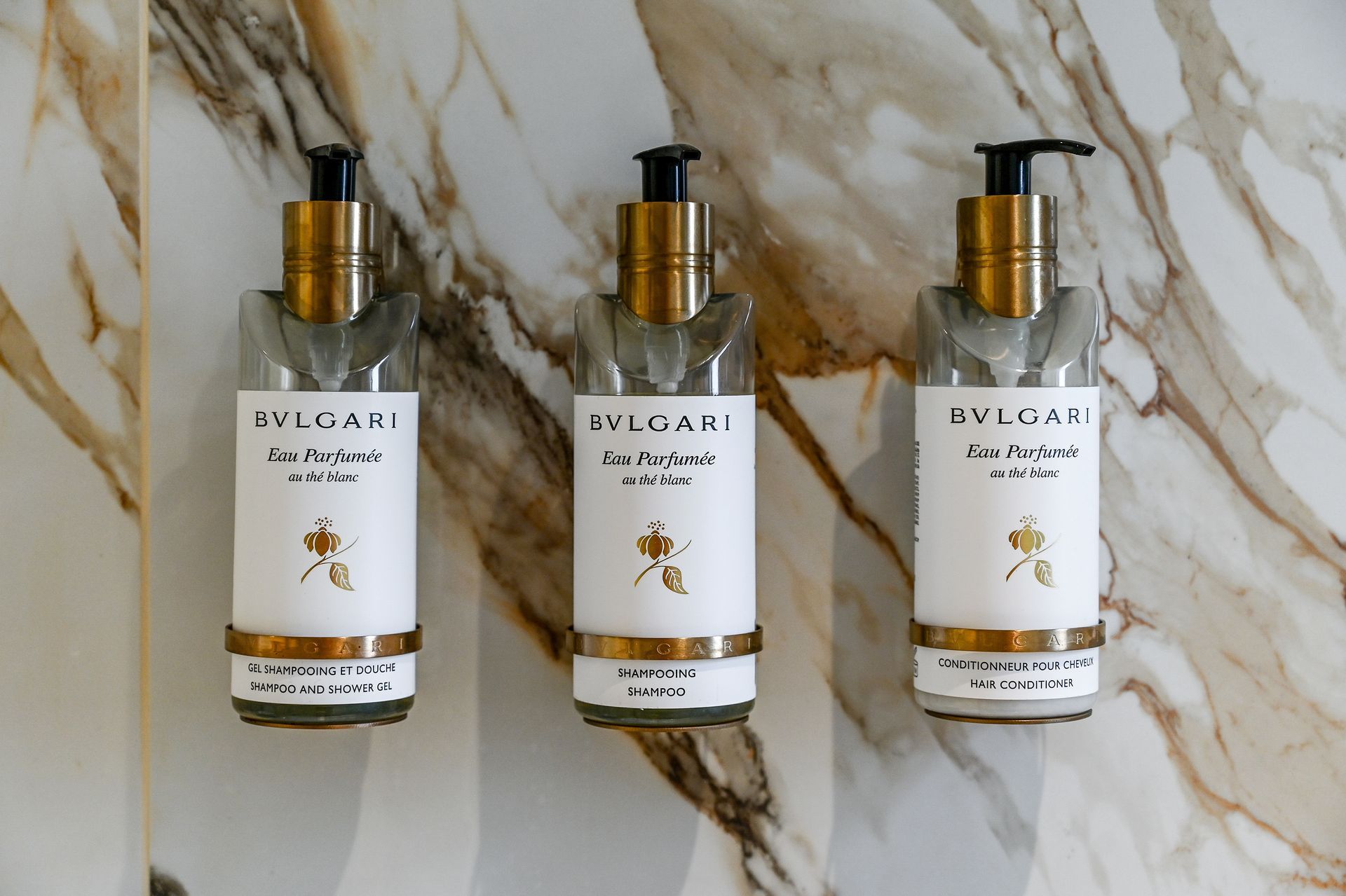 Three gold-pump cosmetic bottles on a marble surface, arranged in a row.