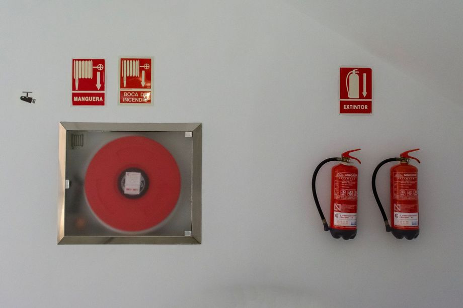 Wall-mounted fire hose reel, extinguisher signs, and two red fire extinguishers on a white wall.