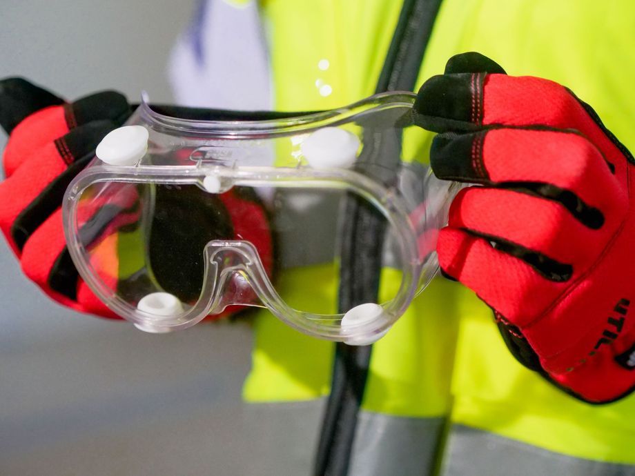 Person holding clear safety goggles with red gloves and a high-vis vest