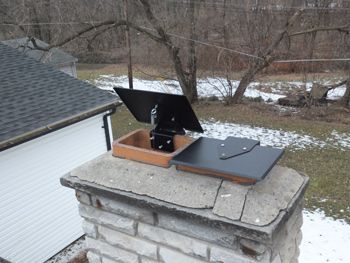 Black metal chimney cap on a stone chimney with snow and bare trees in the background