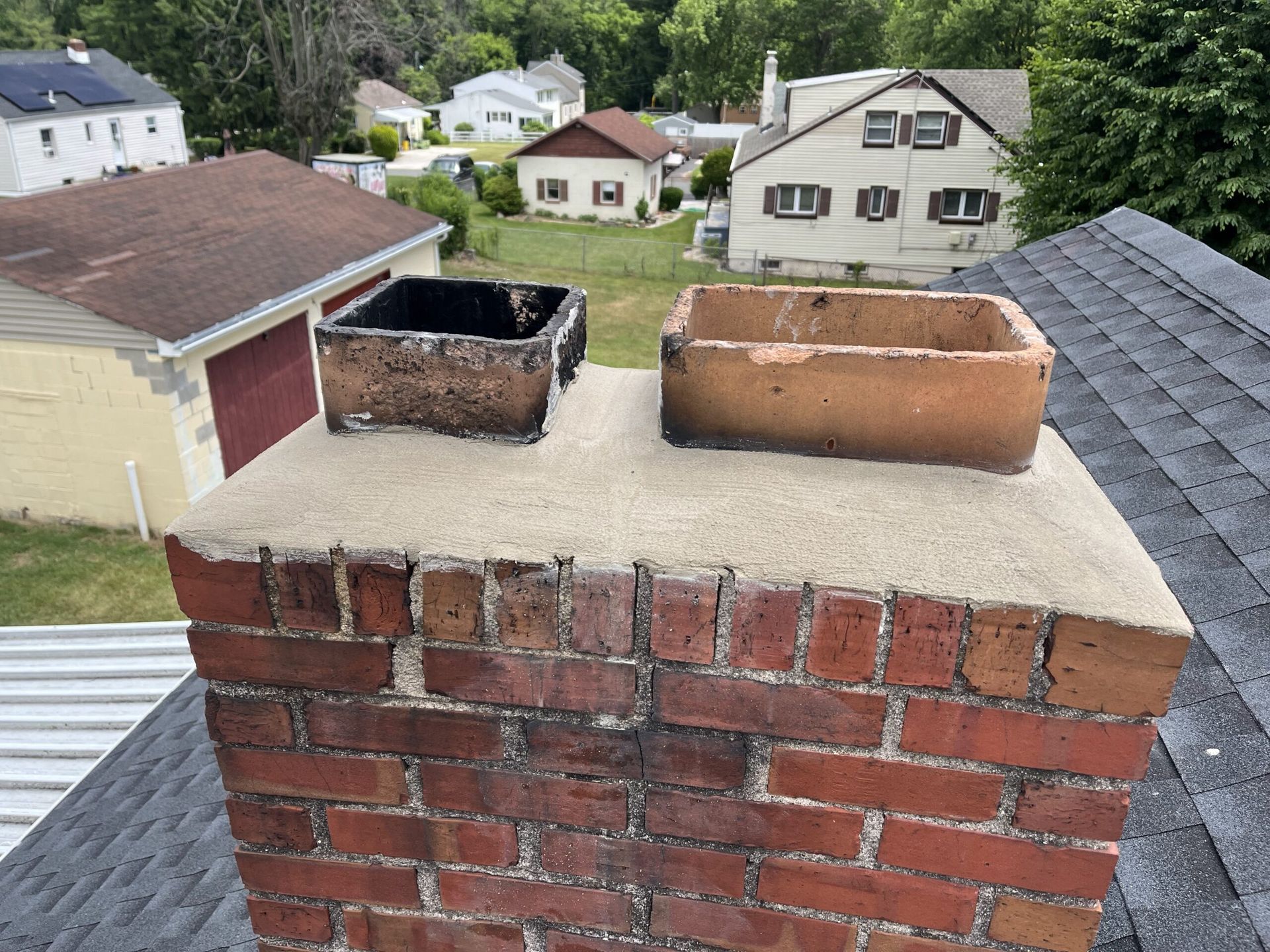 Red brick chimney with two open flue liners on a shingled roof, overlooking a suburban neighborhood.
