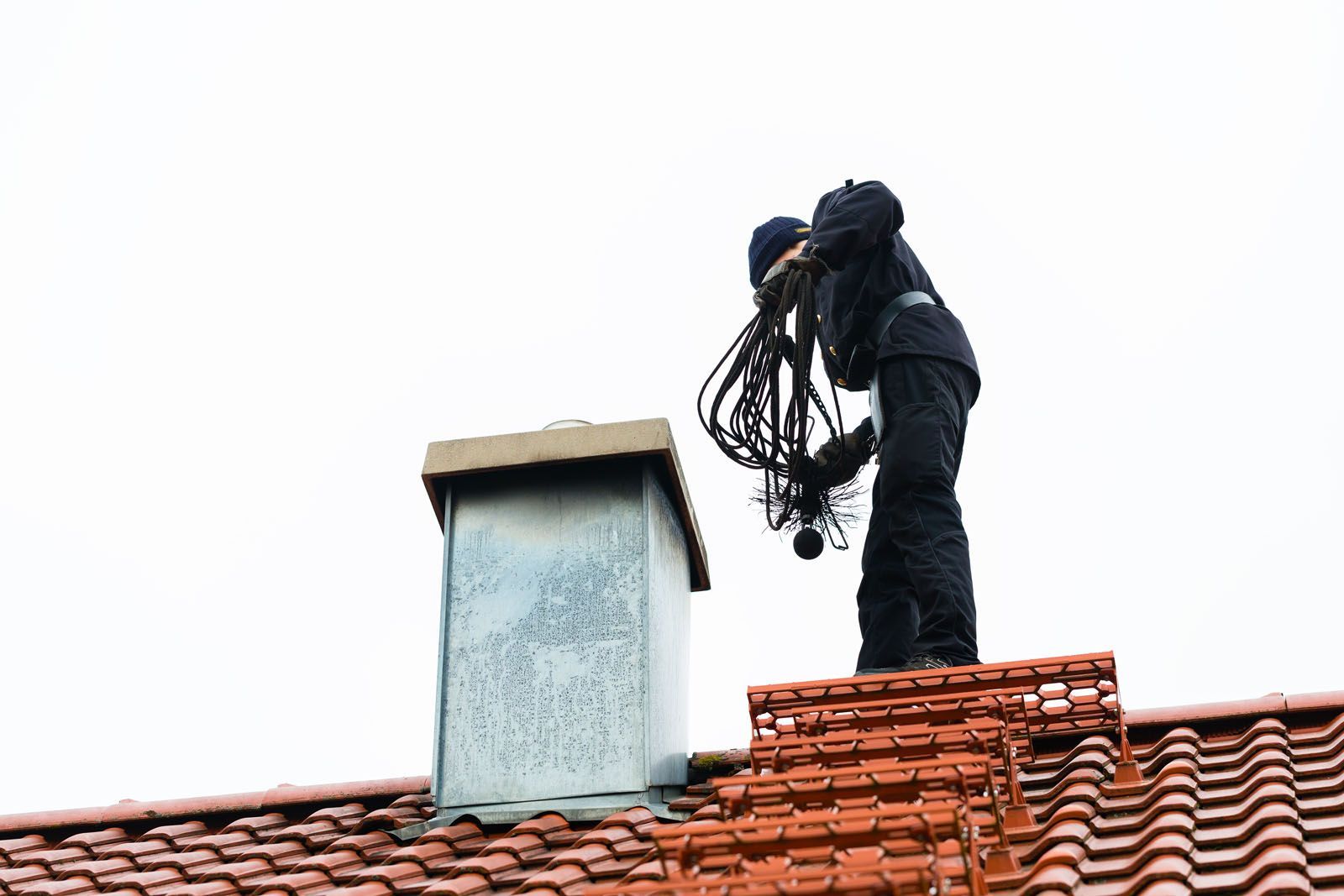 Worker on a red-tiled roof handling cables beside a chimney.