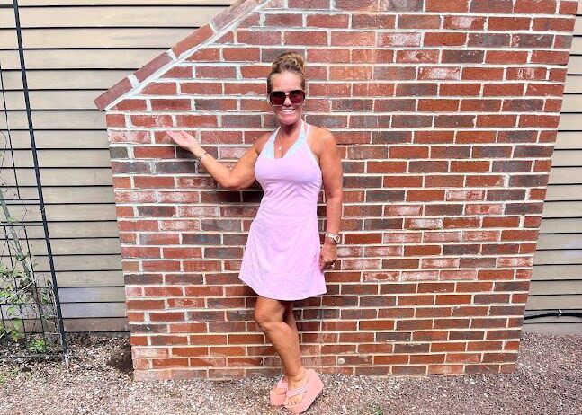 Woman in a lavender dress and sunglasses posing by a brick wall outdoors.