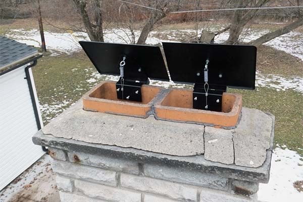 Two black chimney flue dampers on a brick rooftop with snow in the yard behind them