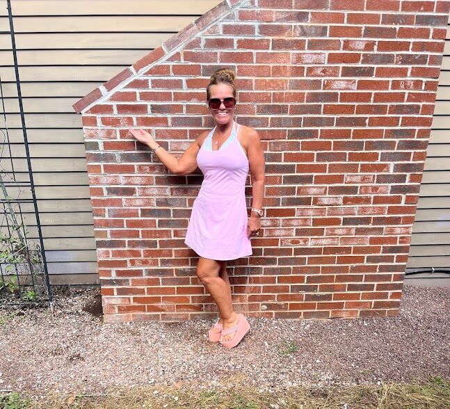 Woman in pink dress and sunglasses posing against a brick wall, pointing upward.