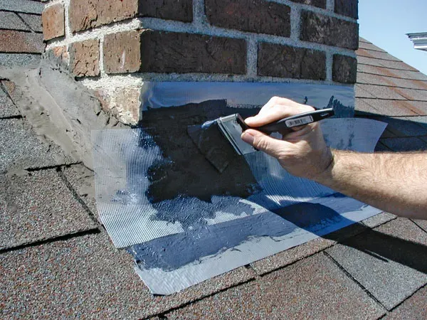 Hand applying black roof sealant around a brick chimney on gray shingles