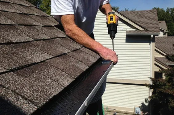 Worker installing a gutter on a shingled roof with a power drill