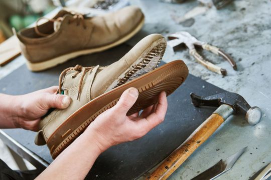Hands repairing a worn beige shoe on a workbench with a hammer nearby