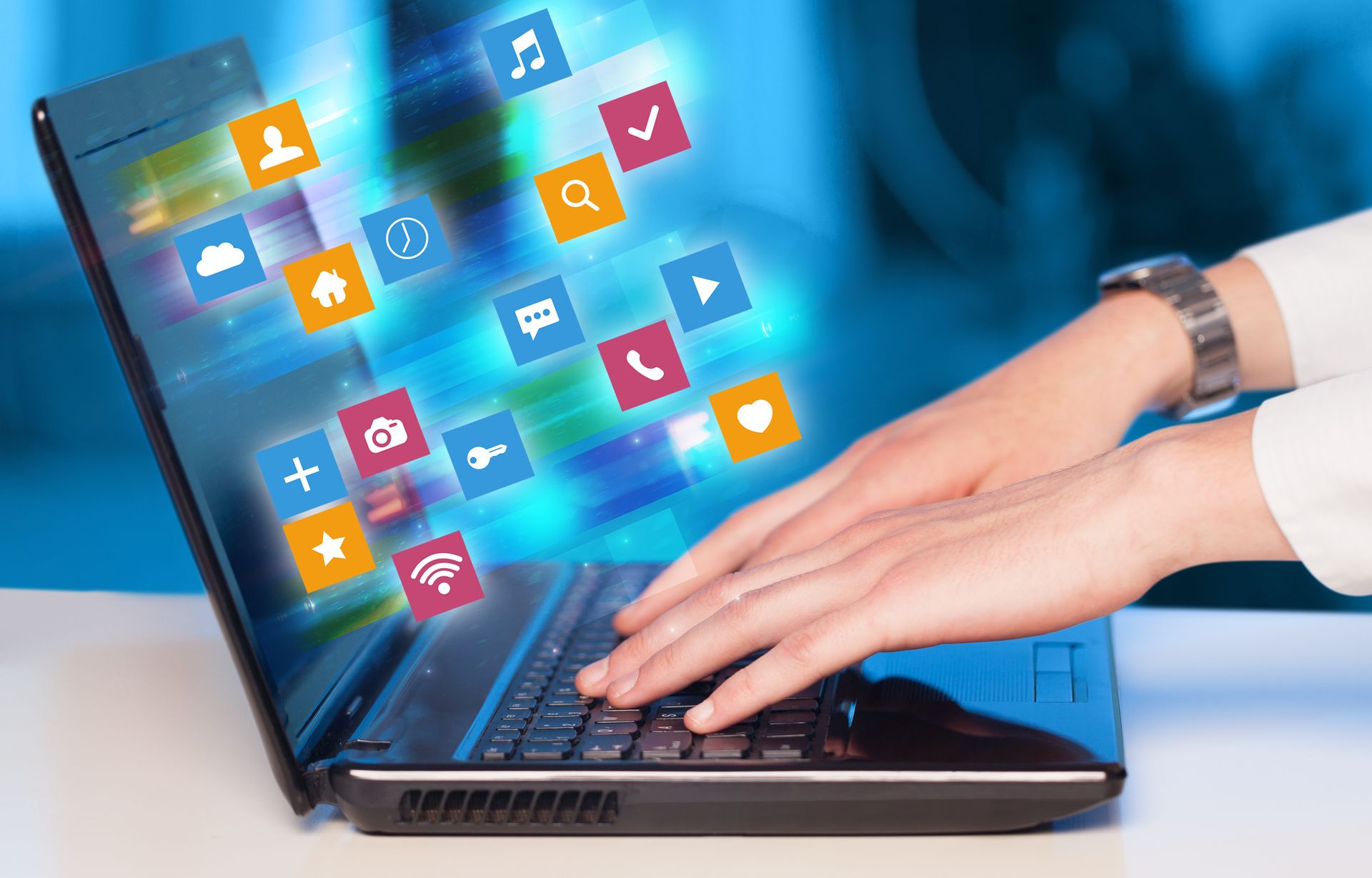 Hands typing on a laptop with colorful app icons floating from the screen against a blurred blue background.