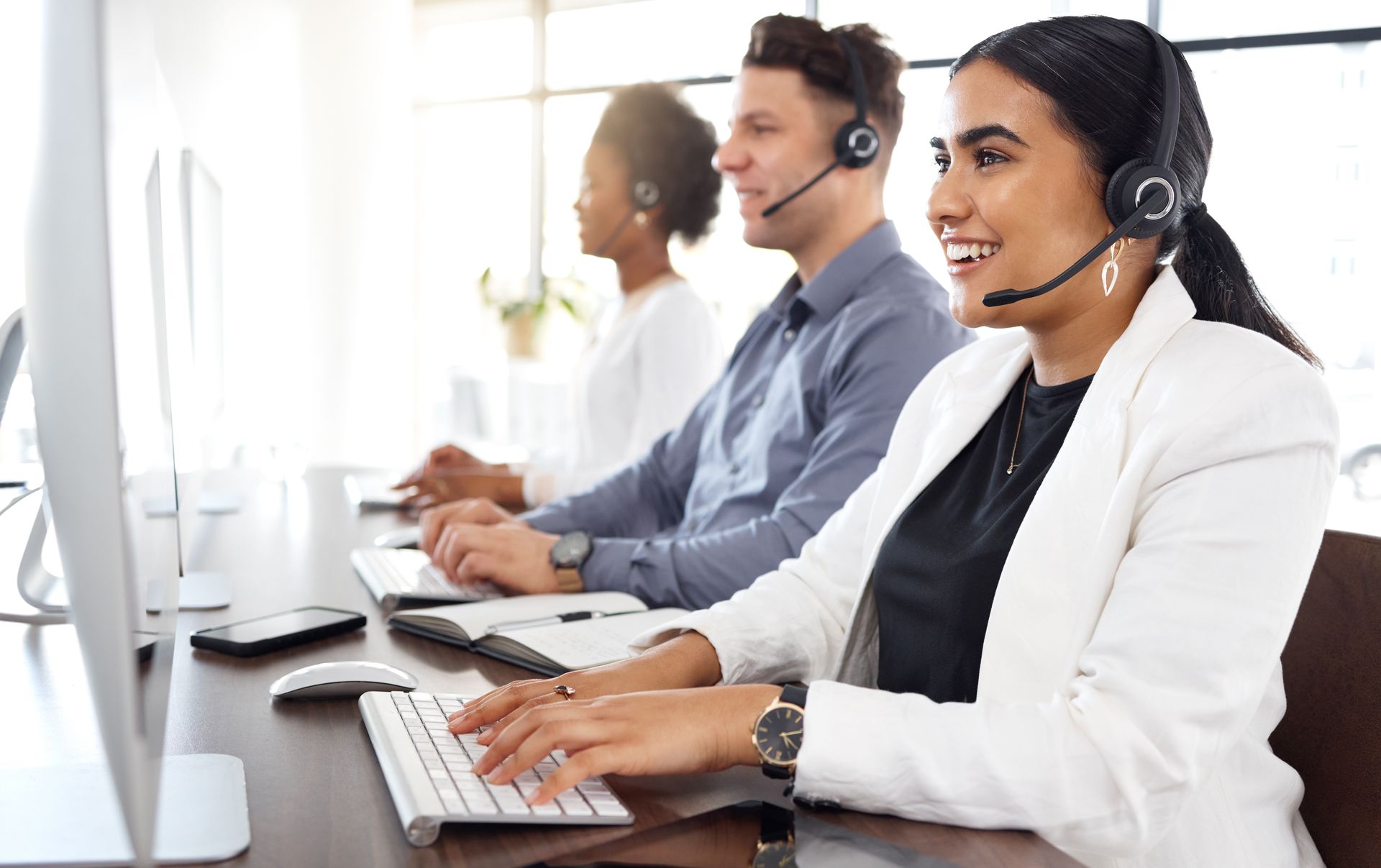 Three customer service representatives, wearing headsets, sit at computers in a brightly lit office, typing and smiling.