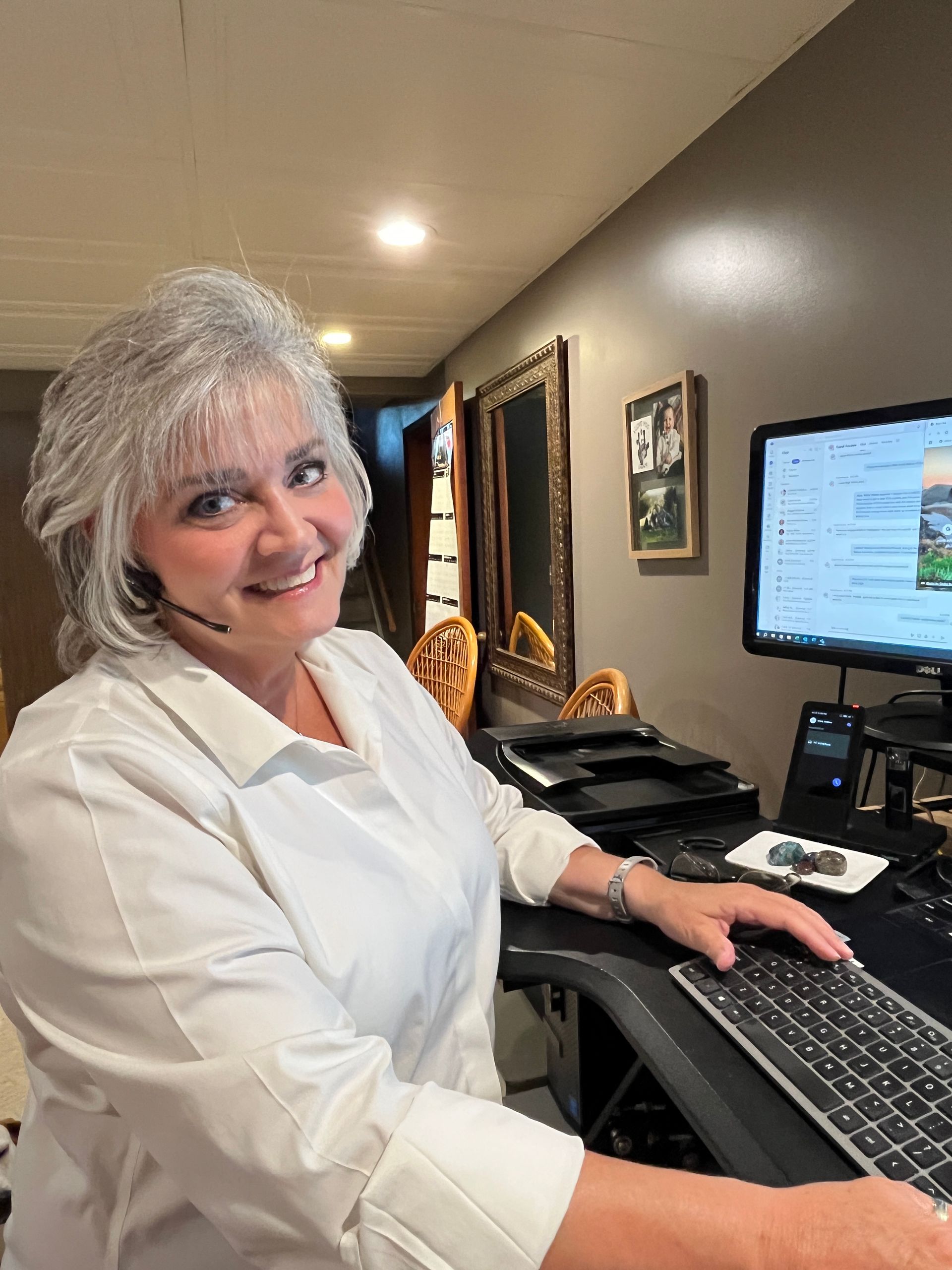 Woman with gray hair smiles while working at a computer. She wears a white shirt in a home office setting.