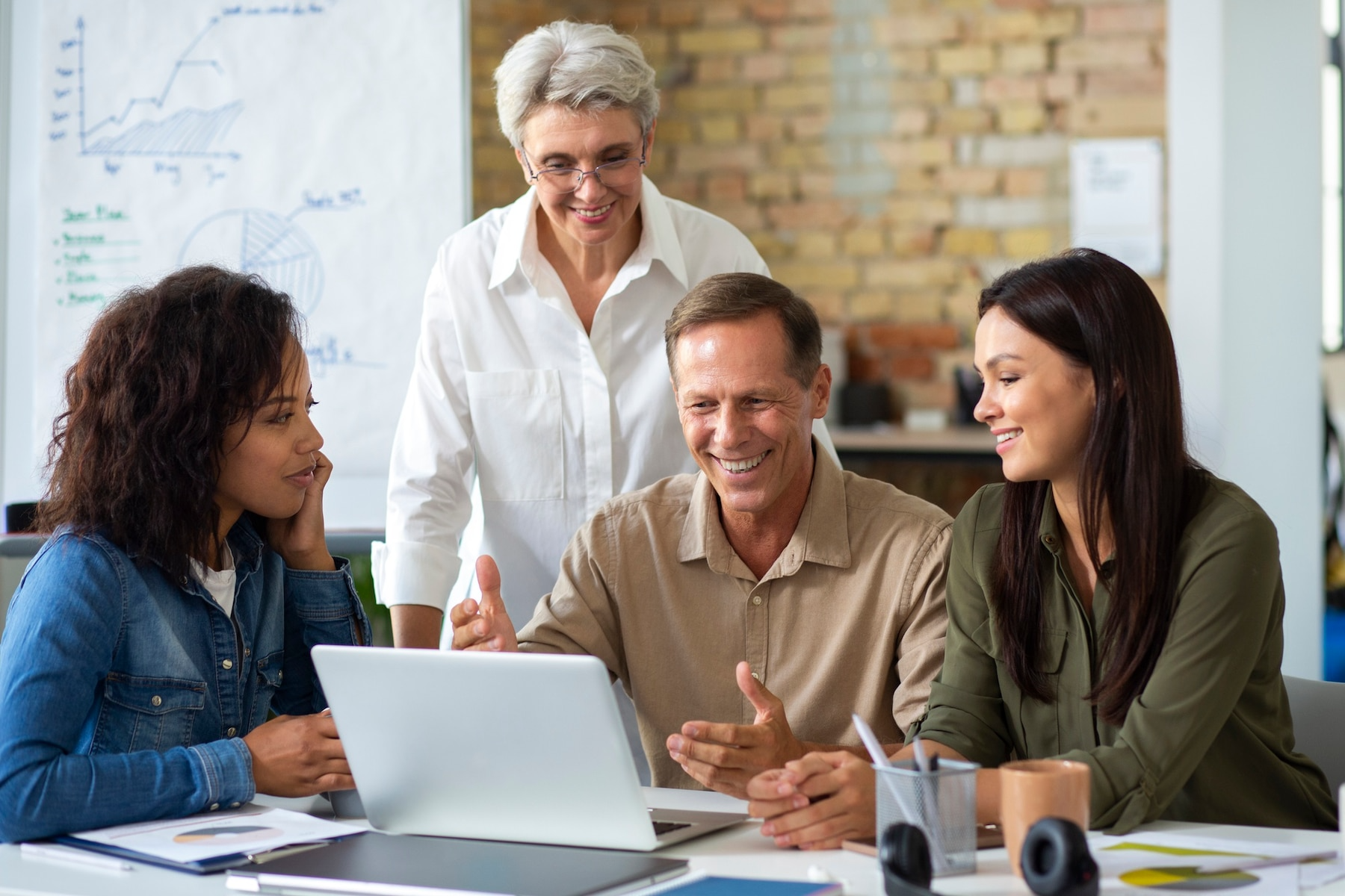 A diverse team smiles while looking at a laptop in a bright office setting; one person stands behind them.