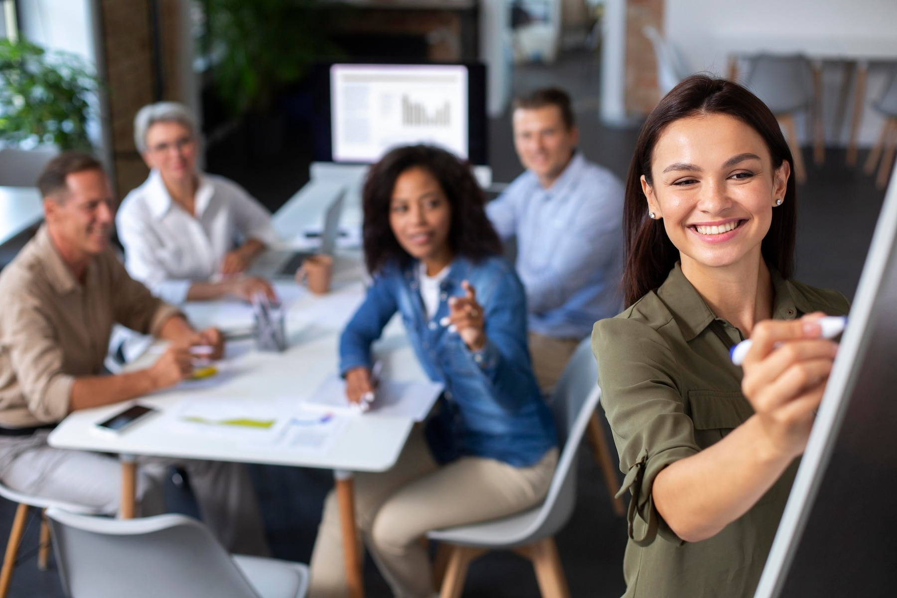 Smiling woman writing on a whiteboard during a meeting, with colleagues seated around a table in a modern office.
