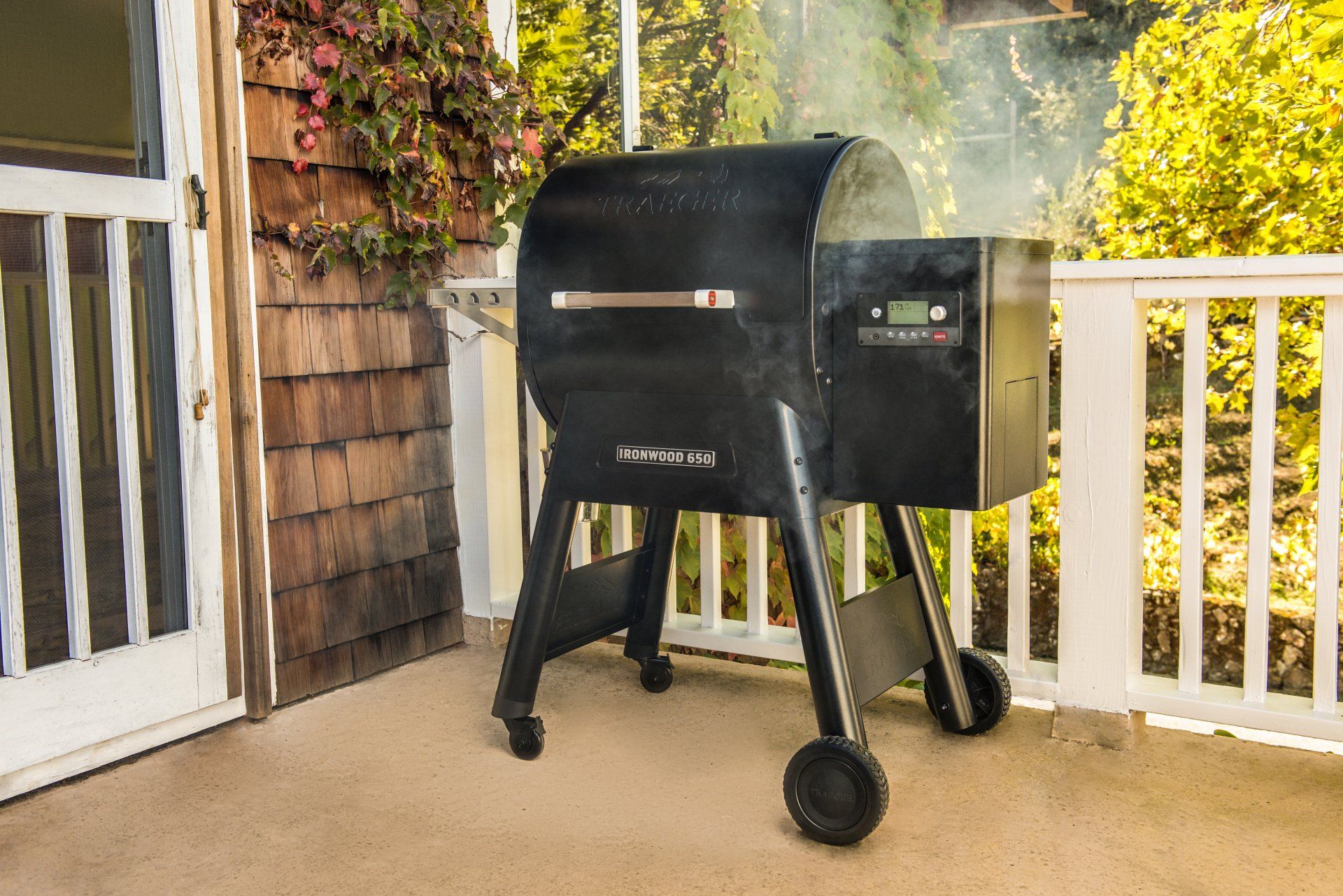 Un barbecue à pellets Traeger noir est installé sur une terrasse, dégageant une légère fumée, à côté d'une maison au toit de bardeaux de bois.