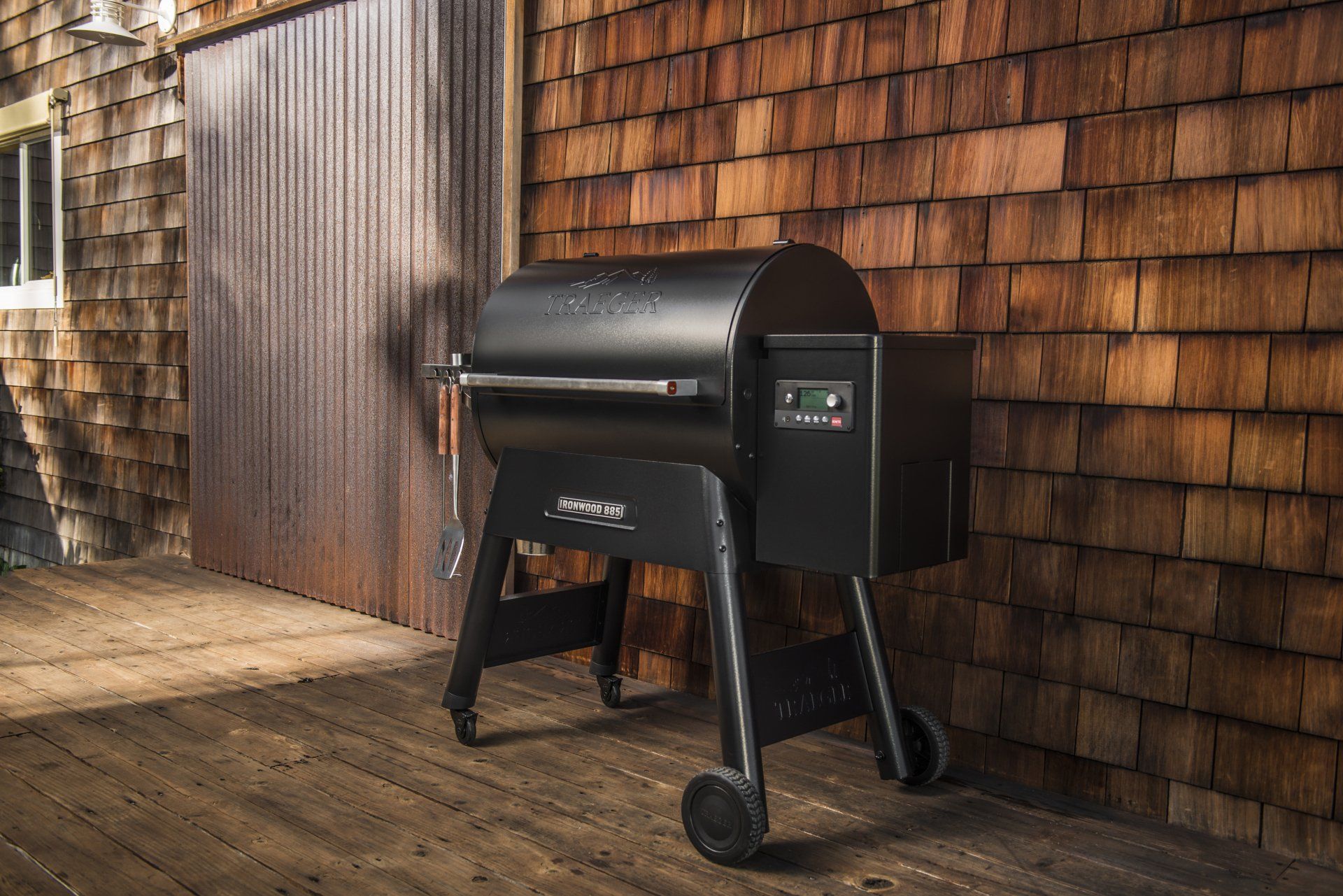 Un barbecue à granulés noirs est installé sur une terrasse en bois, devant un mur recouvert de bardeaux et une porte coulissante en métal.